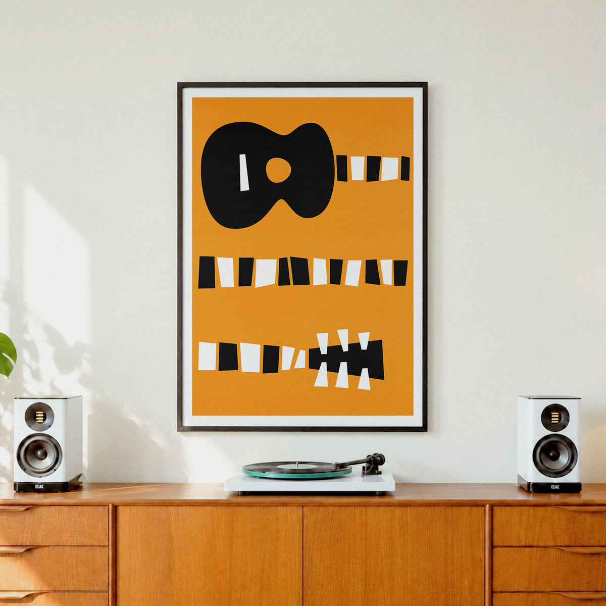 A framed abstract art print featuring a black guitar silhouette and striped shapes on a mustard yellow background, displayed on a wooden sideboard in a modern living room setting with speakers and a turntable.