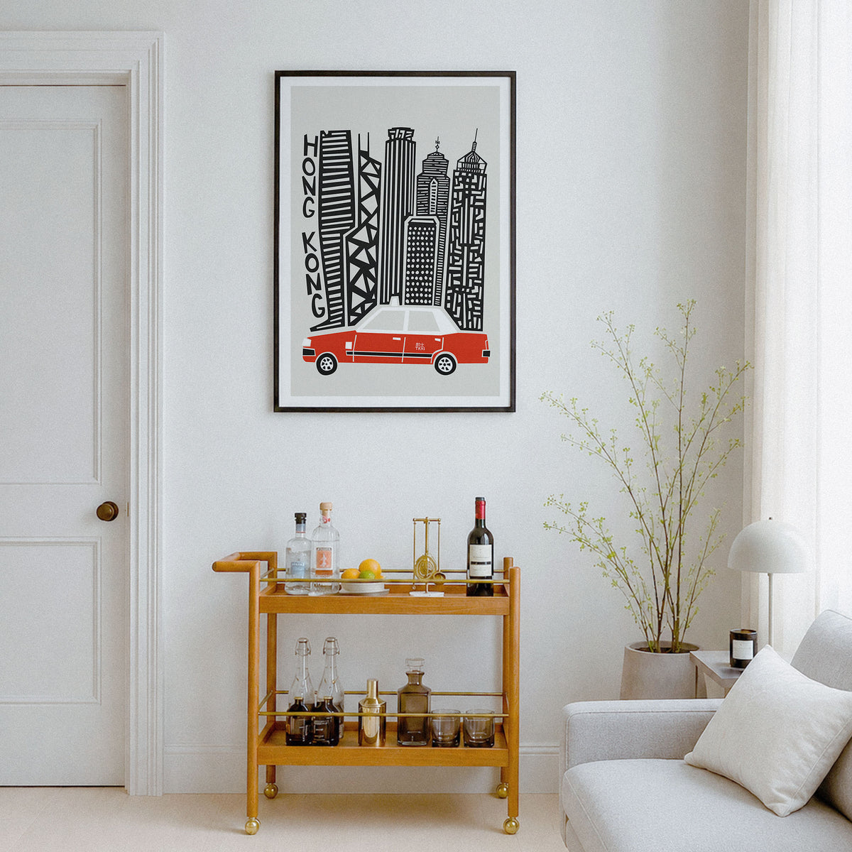 A framed art print featuring a black and white stylized cityscape of Hong Kong with a red car, displayed on a white wall in a modern living room setting above a wooden bar cart.