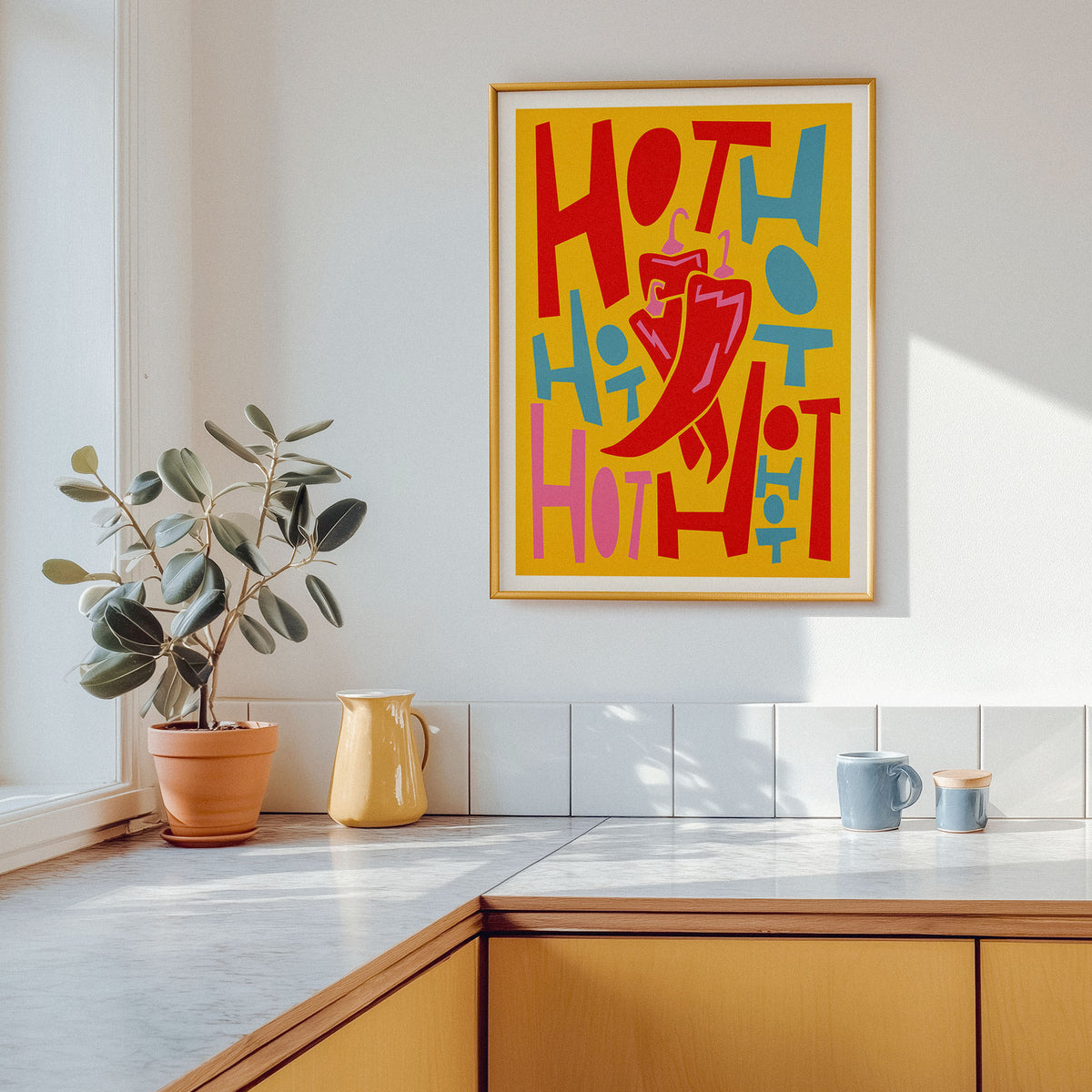 A colorful food art print of some chilli's, featuring the word 'HOT' in bold red, blue, and pink lettering on a yellow background, hanging on a white wall above a kitchen counter with a potted plant and mugs.