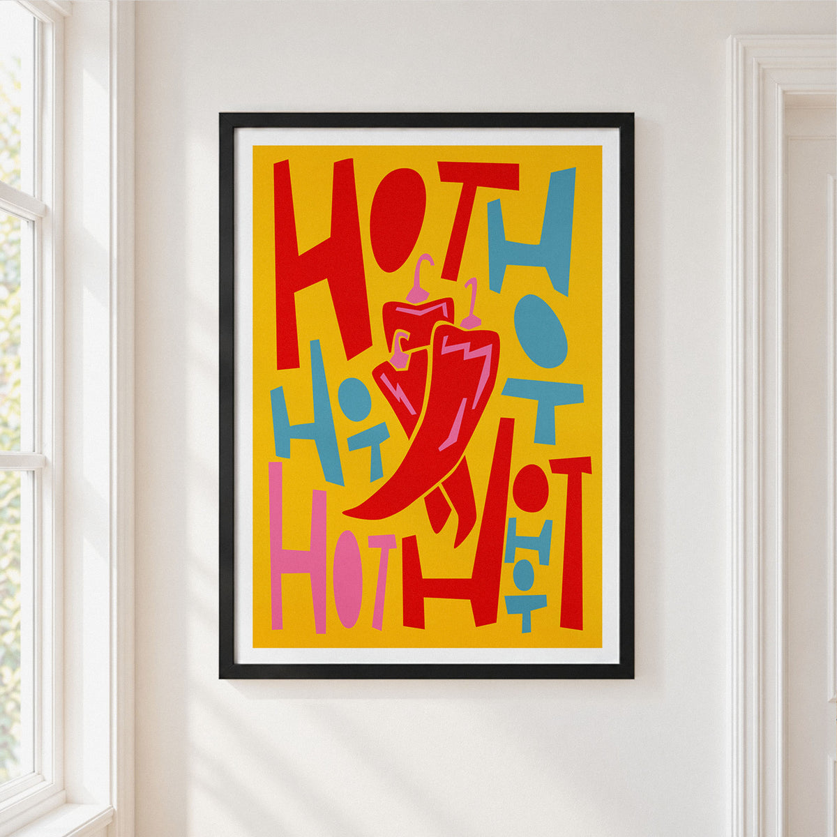 A framed chilli's art print featuring a bold, retro-style graphic with the word 'HOT' repeated in red, pink, and blue on a yellow background, with a large red chili pepper in the center. The print is framed in black with a white mat, hanging on a white wall next to a window.