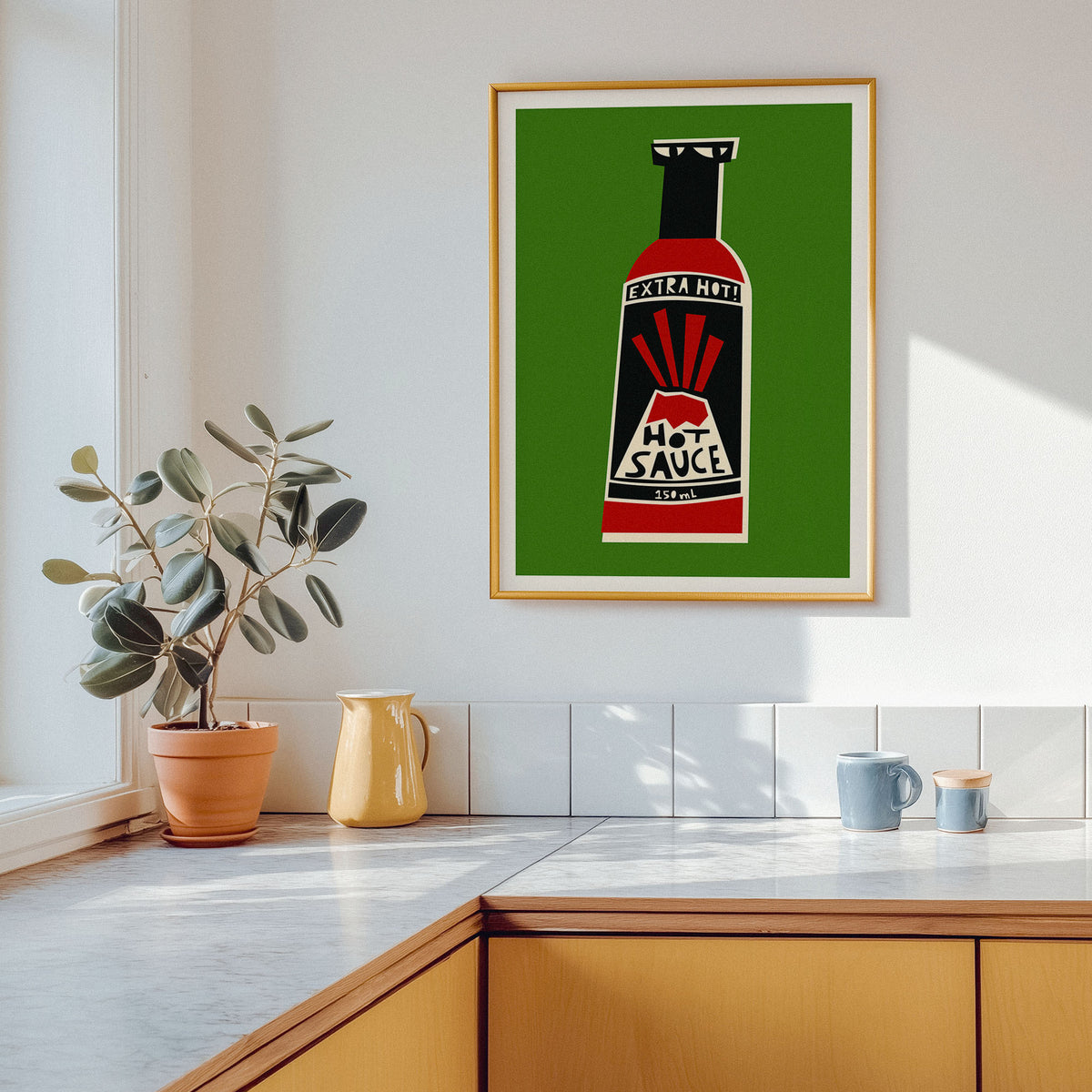 A framed art print featuring a retro hot sauce bottle on a green background, hanging on a white kitchen wall above a wooden counter with a potted plant and mugs.