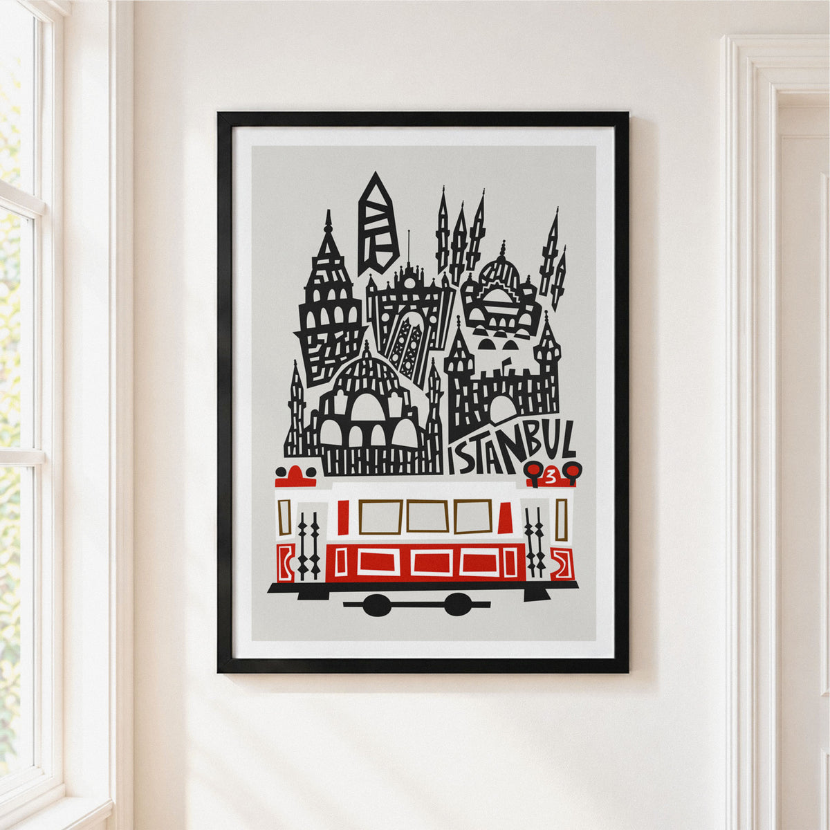 A framed art print featuring a stylized black and red Istanbul skyline with a tram and the word 'ISTANBUL', displayed on a light beige wall next to a window.