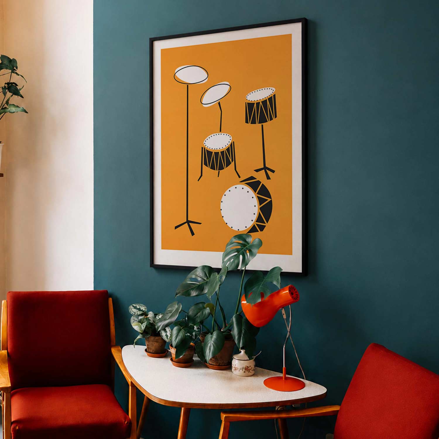 A framed wall art print featuring a minimalist black and white drum set illustration on a mustard yellow background, displayed on a dark teal wall above a small table with potted plants and red chairs.