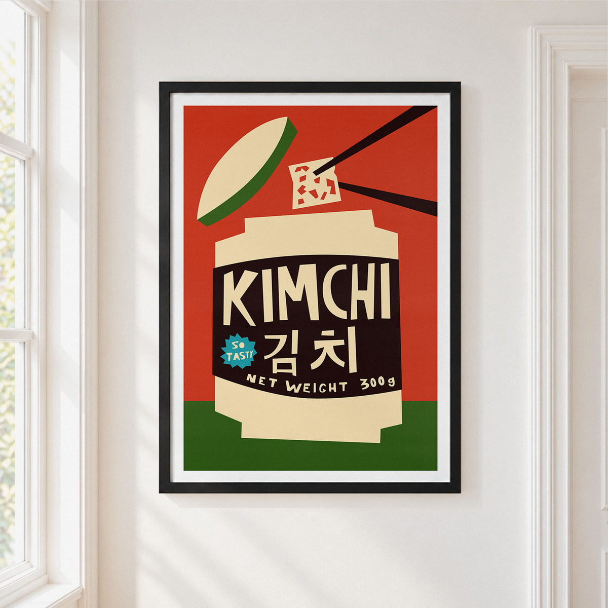 A framed art print featuring a retro-style illustration of a kimchi jar with the text 'KIMCHI 김치' and 'NET WEIGHT 300g', hanging on a white wall next to a window.