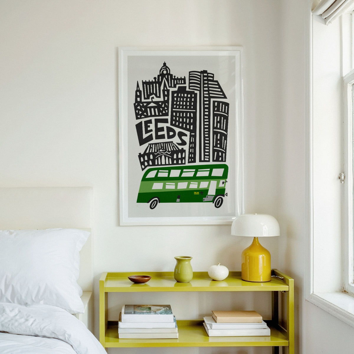 A framed art print featuring a black and white city skyline with a green double-decker bus and the word 'LEEDS' on a light gray wall above a yellow bedside table in a bedroom setting.