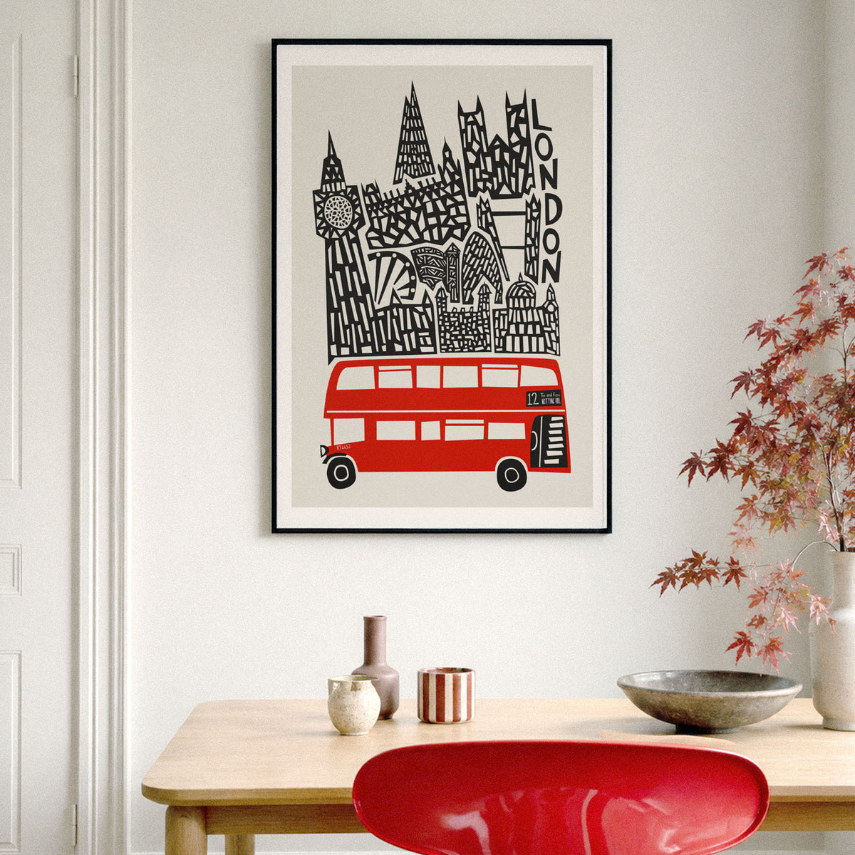 A framed art print featuring a stylized black-and-white London skyline with a red double-decker bus, hanging on a light gray wall above a wooden table with decorative vases, a bowl, and a red chair.