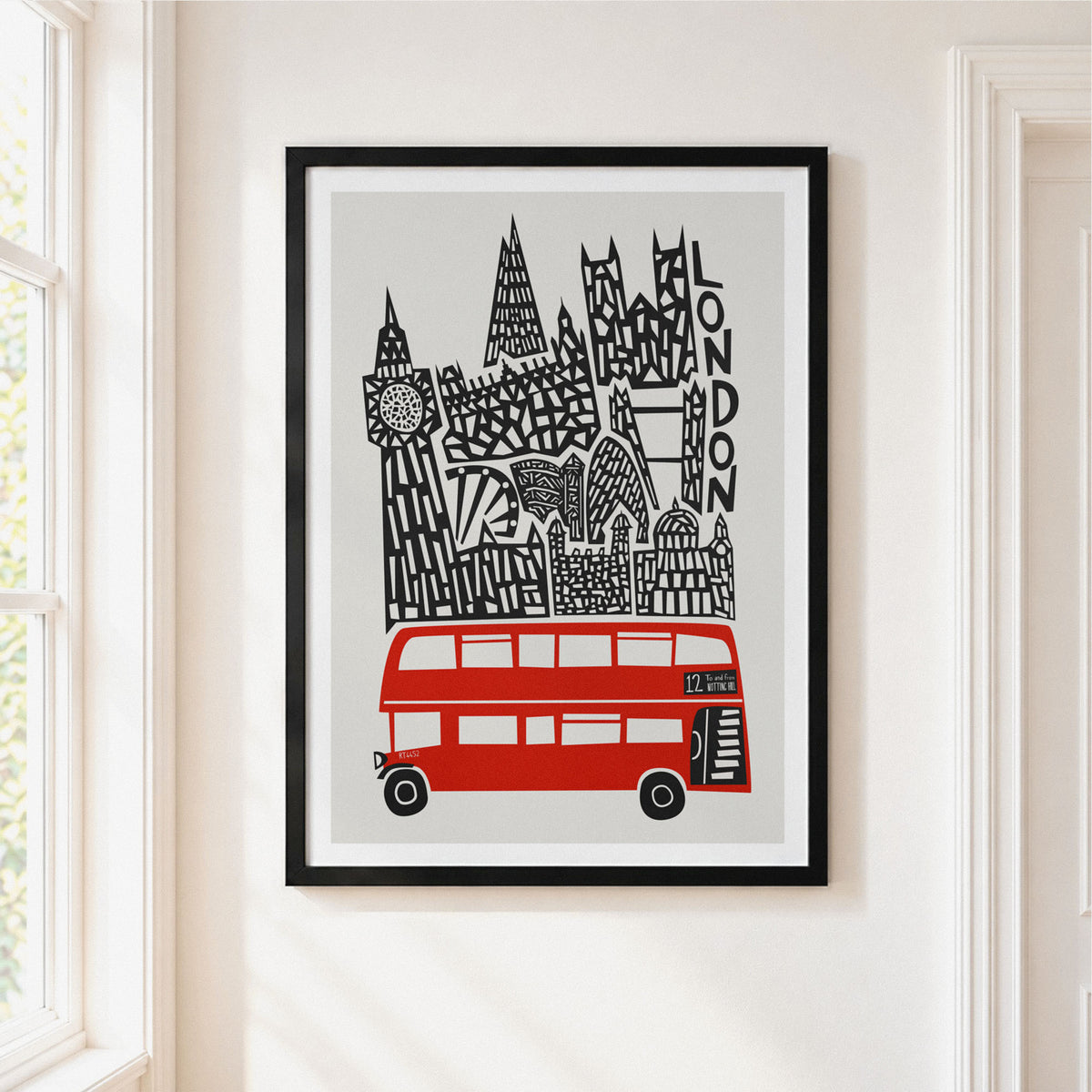 A framed art print featuring a stylized London skyline with a red double-decker bus and black architectural details on a light gray background, hung on a cream-colored wall next to a window.