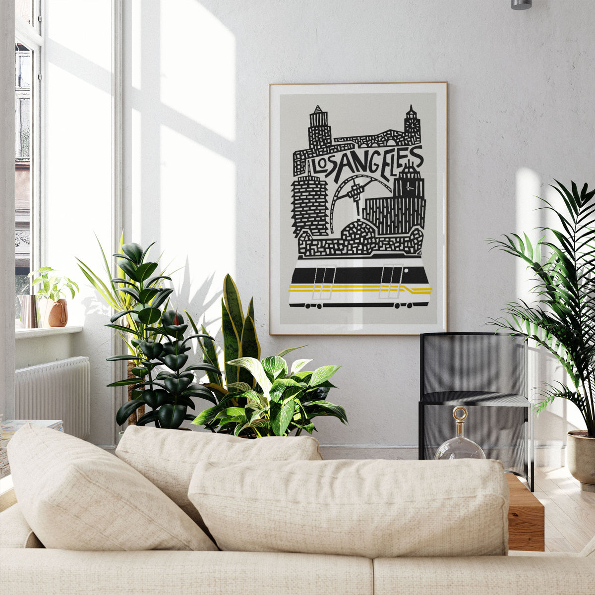 A framed Los Angeles cityscape art print with black and white illustration and a yellow stripe, displayed on a light gray wall above a beige sofa in a sunlit living room with potted plants.