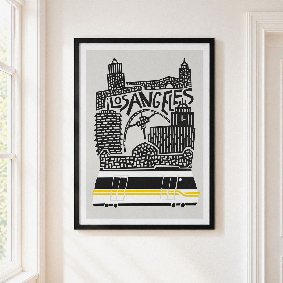 A framed Los Angeles cityscape art print hangs on a white wall next to a window. The print features black silhouettes of the skyline and a train on a light gray background, enclosed in a black frame with a white mat.