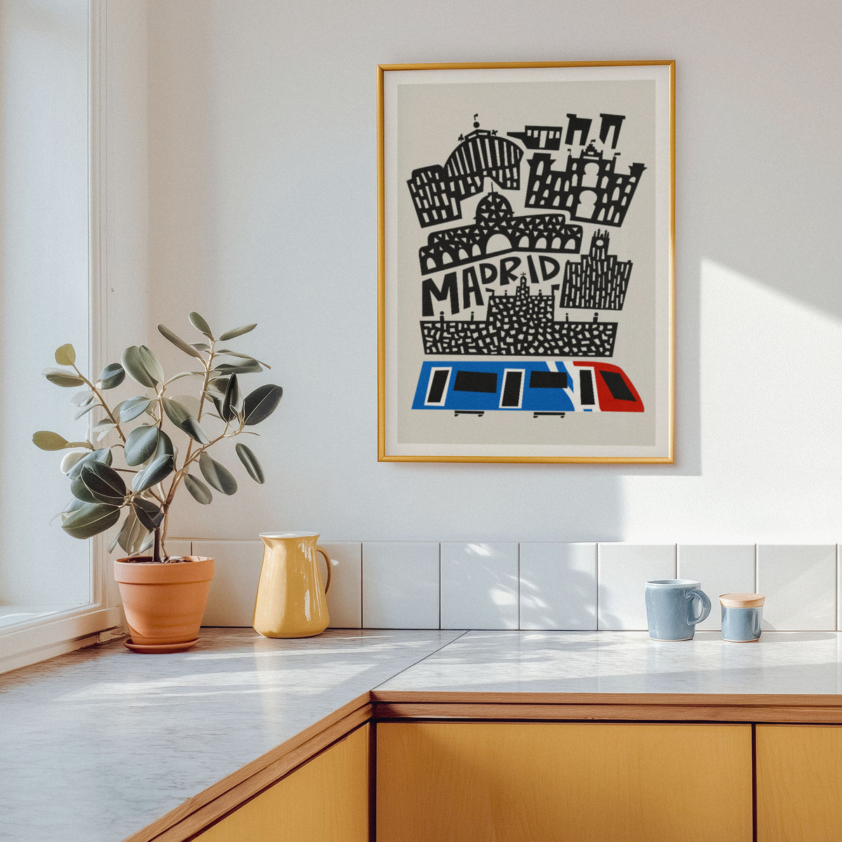 A framed art print featuring a stylized black and white Madrid skyline with a blue and red train, hanging on a white wall above a kitchen counter with a potted plant and mugs.