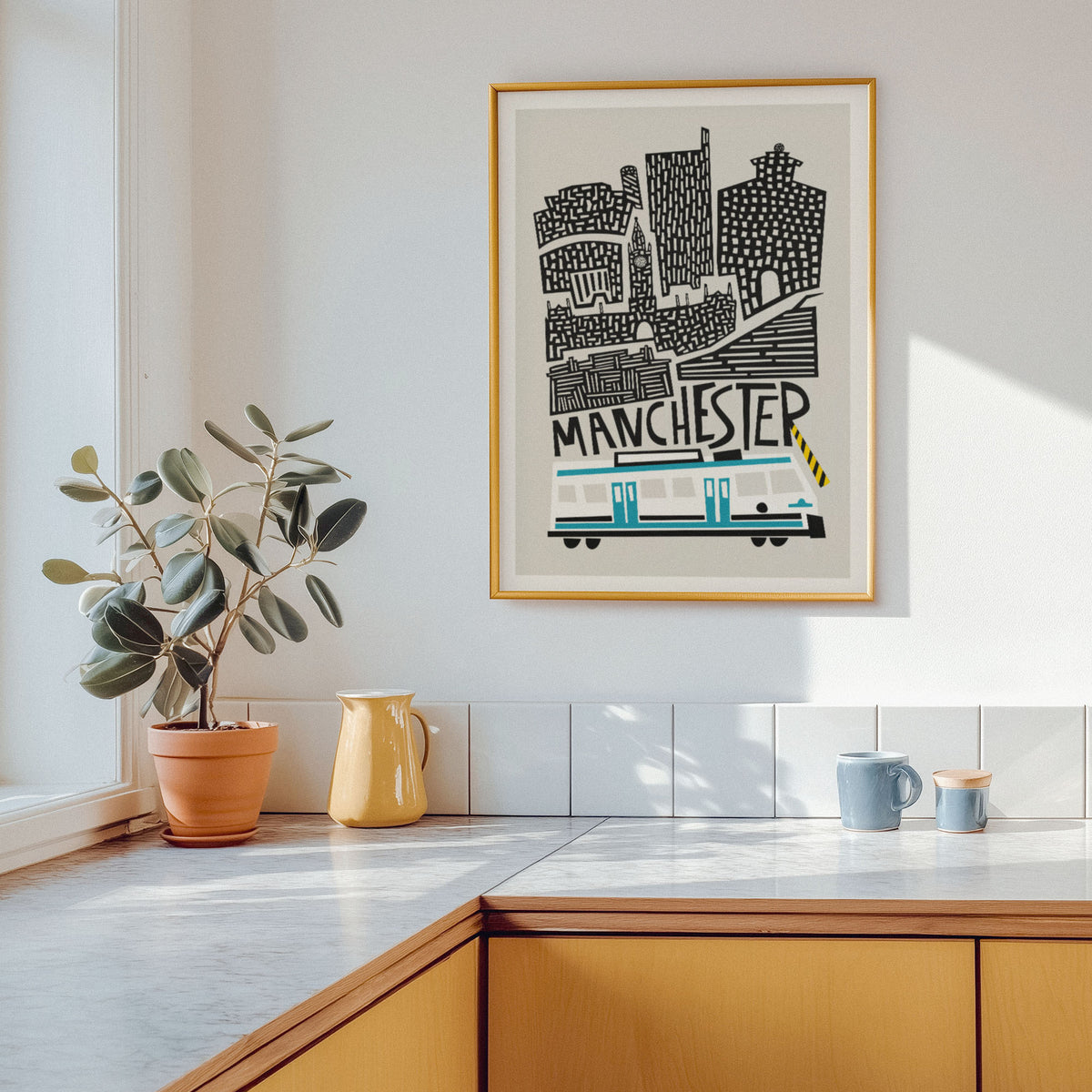 A framed art print featuring a stylized black and white cityscape of Manchester with a tram, hanging on a white kitchen wall above a countertop with a potted plant and ceramic vessels.