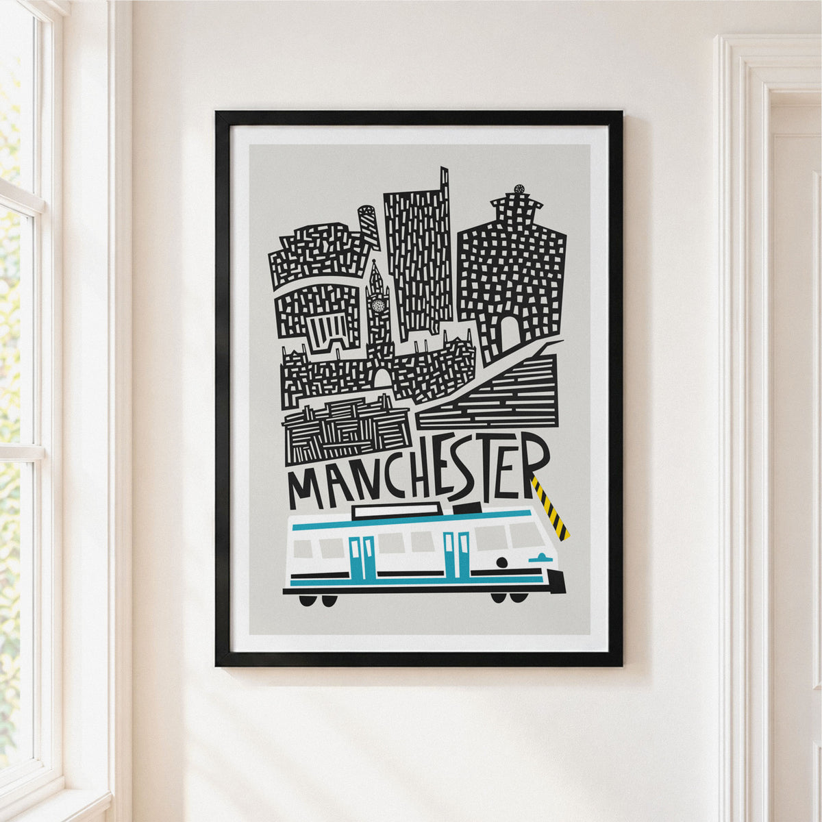 A framed art print featuring a stylized Manchester skyline with a white and grey tram and black architectural details on a light gray background, hung on a cream-colored wall next to a window.
