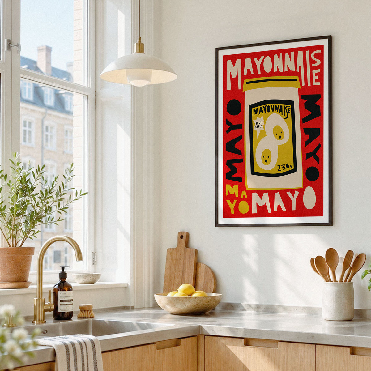 A framed pop-art style poster featuring a yellow mayonnaise can with red, black, and white typography, hanging on a white kitchen wall above a countertop with a sink, plants, and utensils.