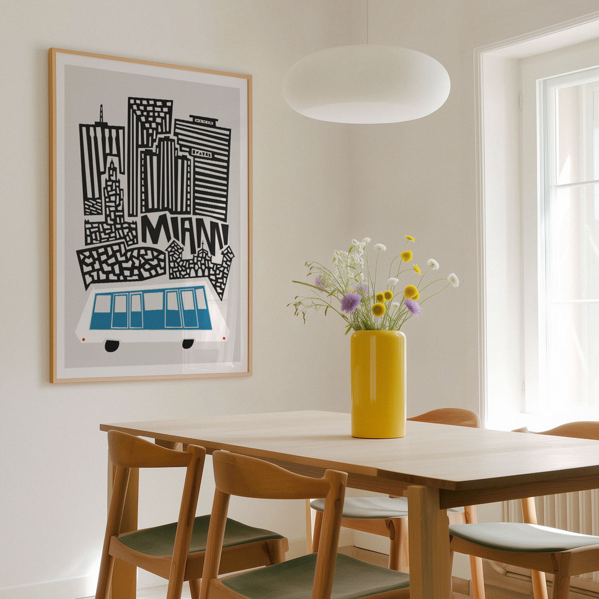A framed art print featuring a stylized black and white city skyline with a blue and white bus and the word 'MIAMI' in a light wood dining room, styled with a yellow vase of flowers on a wooden table.