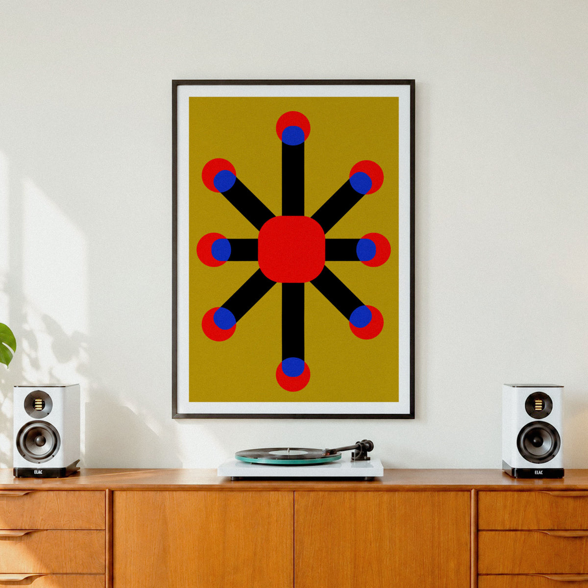 A framed abstract geometric art print featuring a red starburst design on a mustard yellow background, styled on a white wall above a wooden credenza with speakers and a turntable.