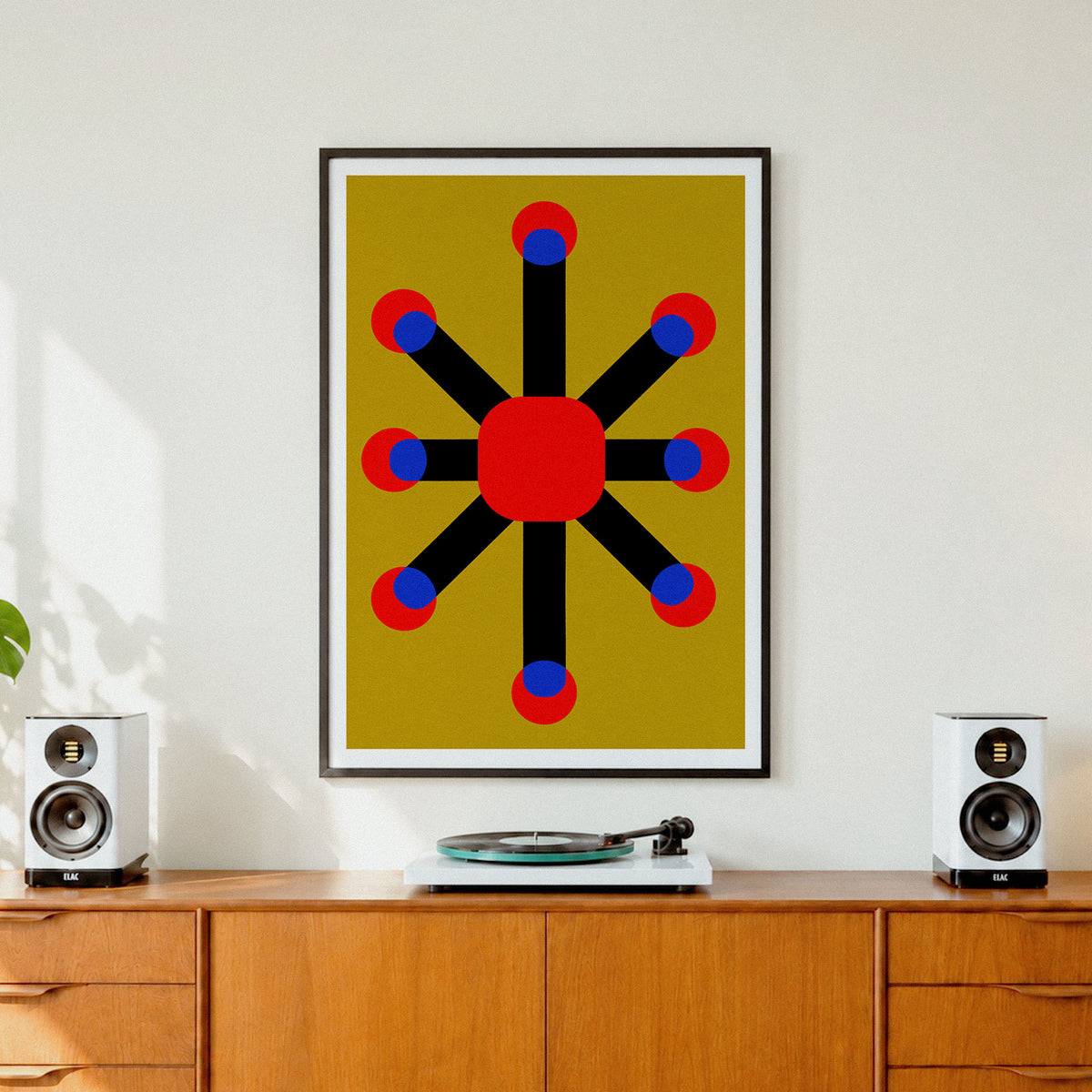 A framed abstract geometric art print featuring a red starburst design on a mustard yellow background, styled on a white wall above a wooden credenza with speakers and a turntable.