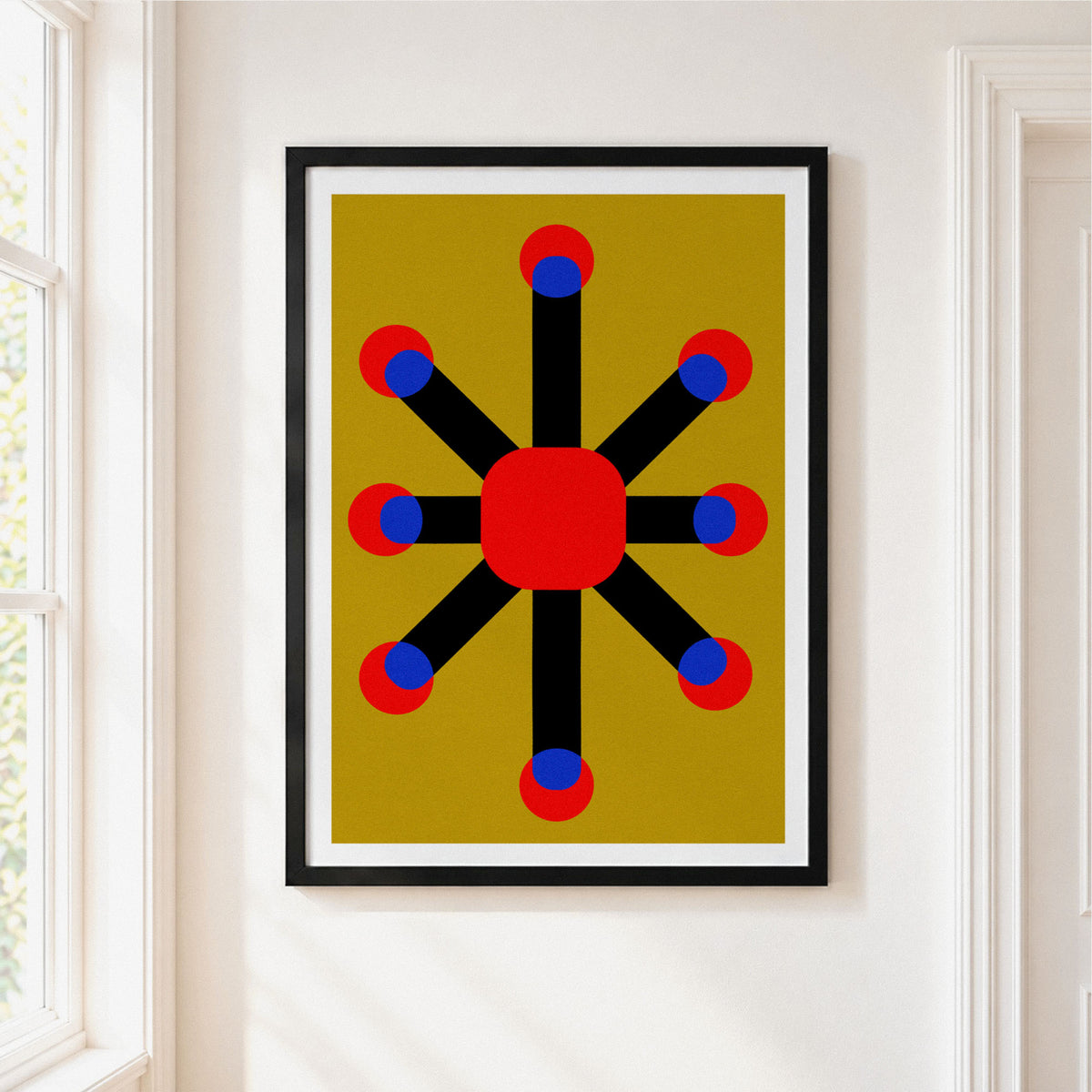 A framed abstract geometric art print hanging on a white wall next to a window. The print features a central red circle connected to eight red and blue circles by black radiating bars, set against a mustard yellow background.
