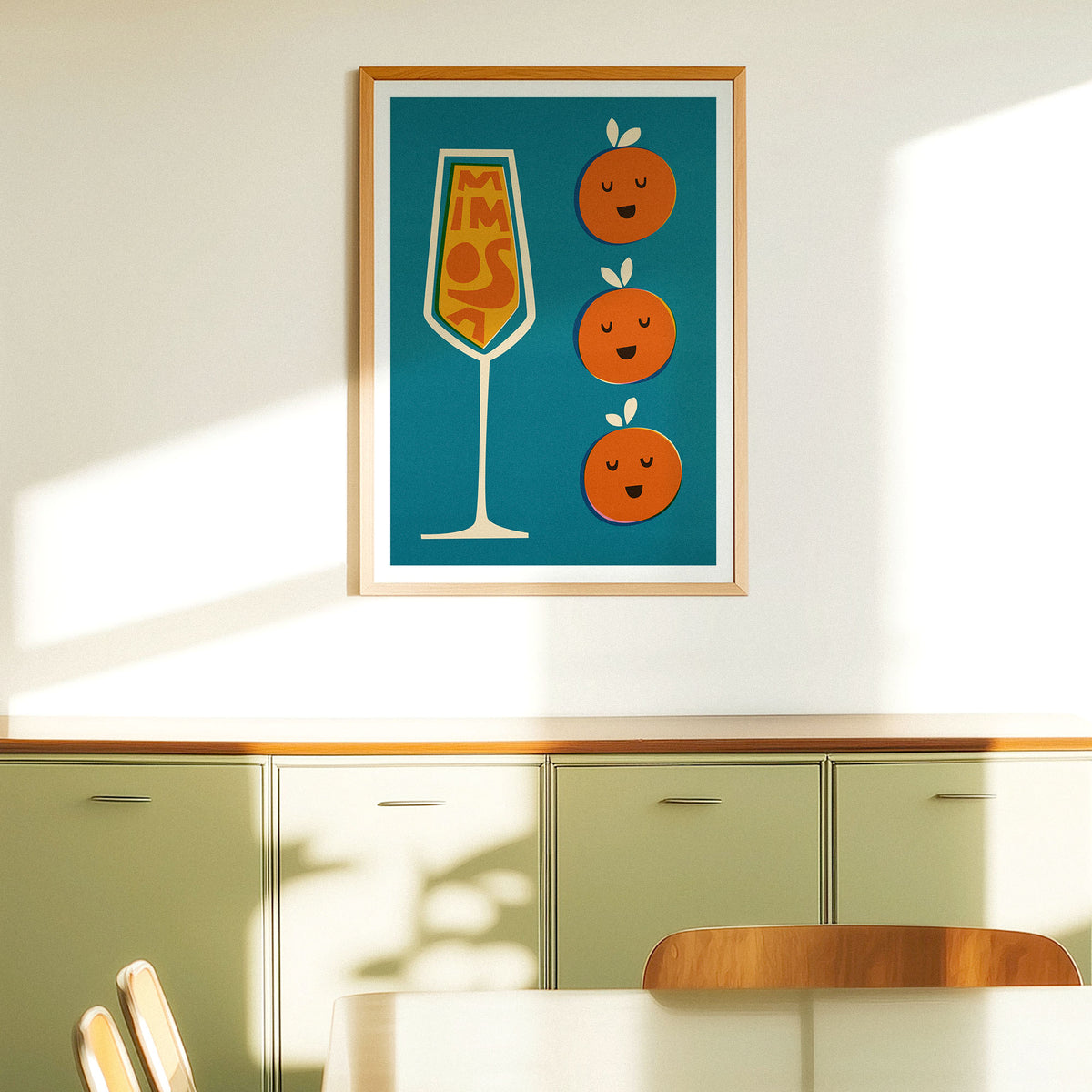 A framed art print featuring a teal background with a stylized champagne flute and three smiling oranges, hung on a white wall above light green kitchen cabinets in a sunlit room.