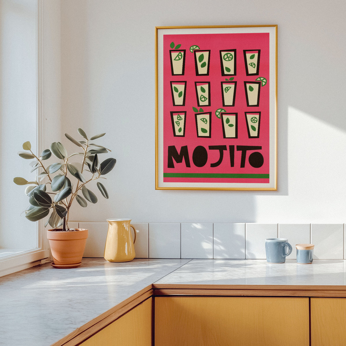 A framed art print featuring a pink background with green and black mojito cocktail illustrations and the word 'MOJITO' at the bottom, hanging on a white wall in a bright kitchen above a wooden counter.