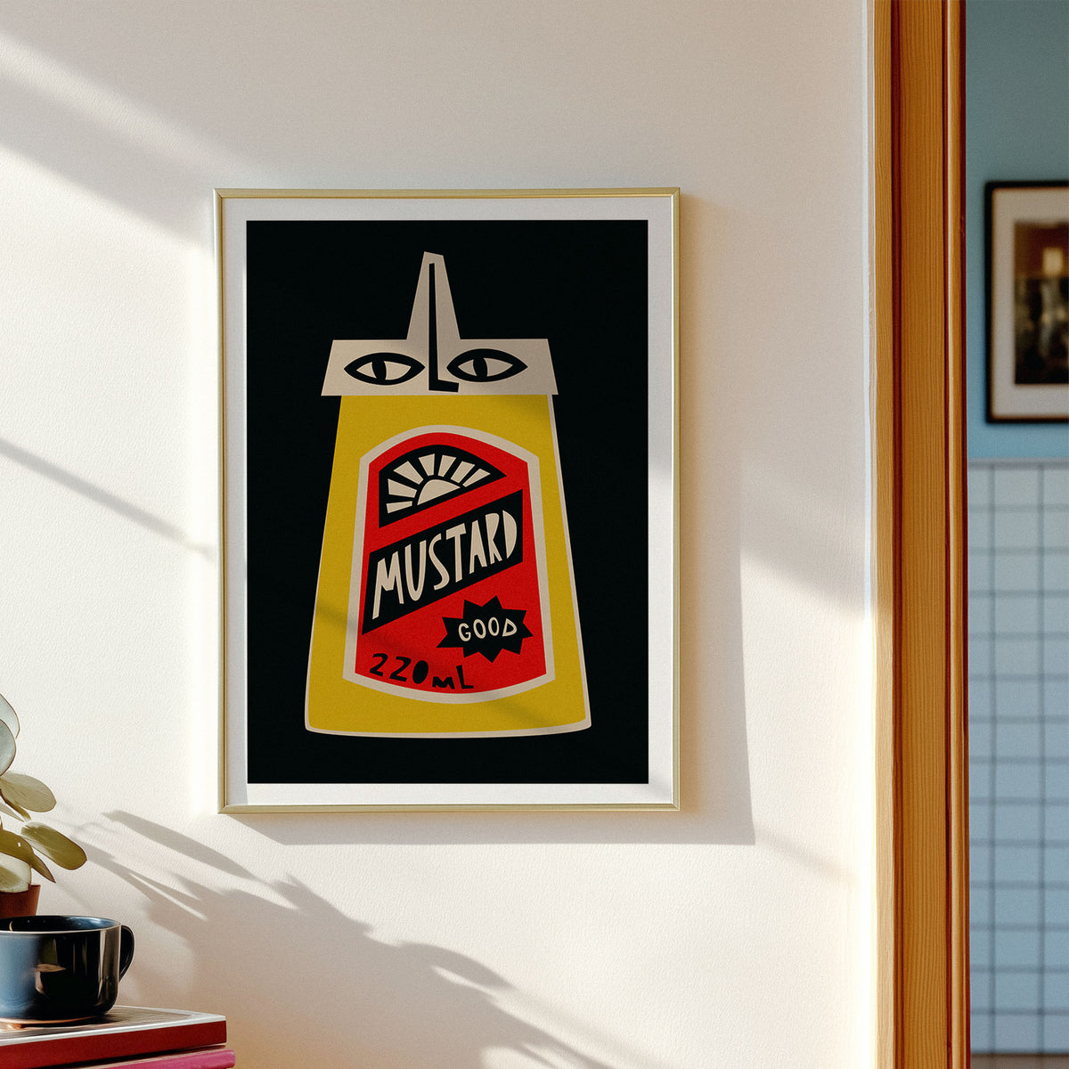 A framed art print featuring a stylized mustard bottle with eyes, hung on a white wall in a sunlit room. The print has a black background with a yellow mustard bottle, red label details, and white text reading 'MUSTARD 220 mL'.