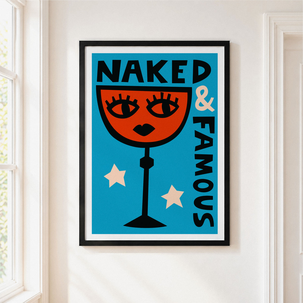 A framed art print featuring a retro-style illustration of a cocktail glass with a female face and the text 'NAKED & FAMOUS' on a bright blue background, hanging on a white wall next to a window.