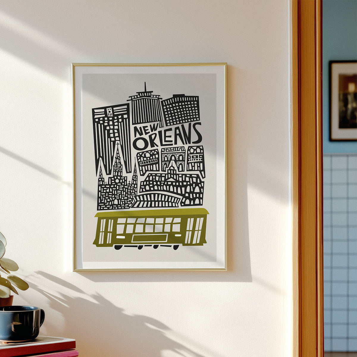 A framed New Orleans cityscape art print hangs on a white wall. The print features black architectural silhouettes and the words 'NEW ORLEANS' above a yellow-green streetcar, set in a light wood frame.