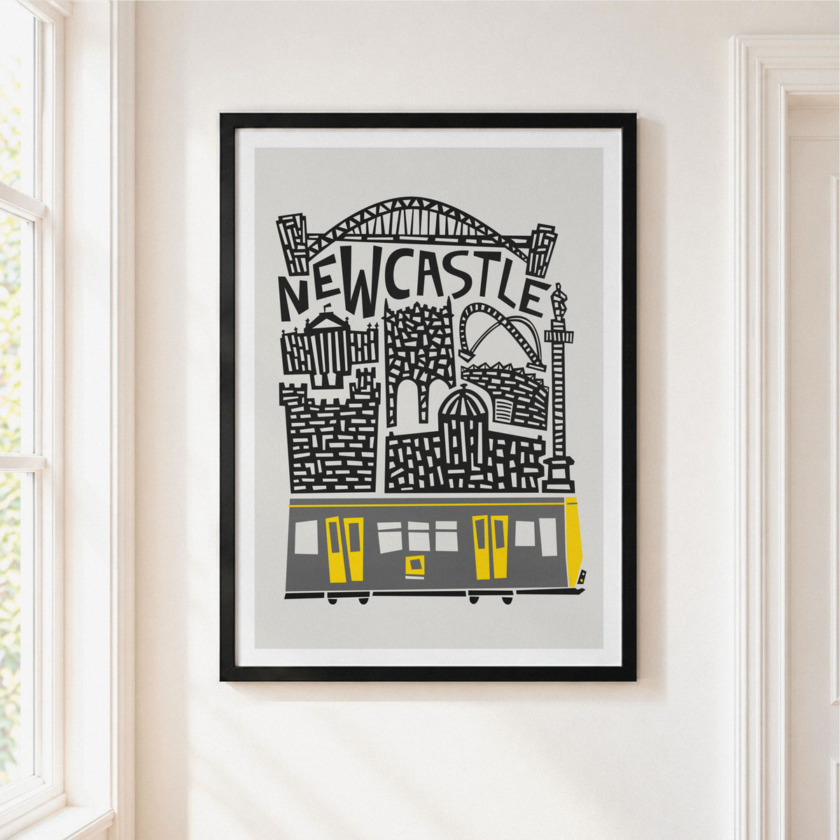 A framed art print featuring a stylized black and gray illustration of Newcastle with a yellow tram, hanging on a white wall in a bright room.