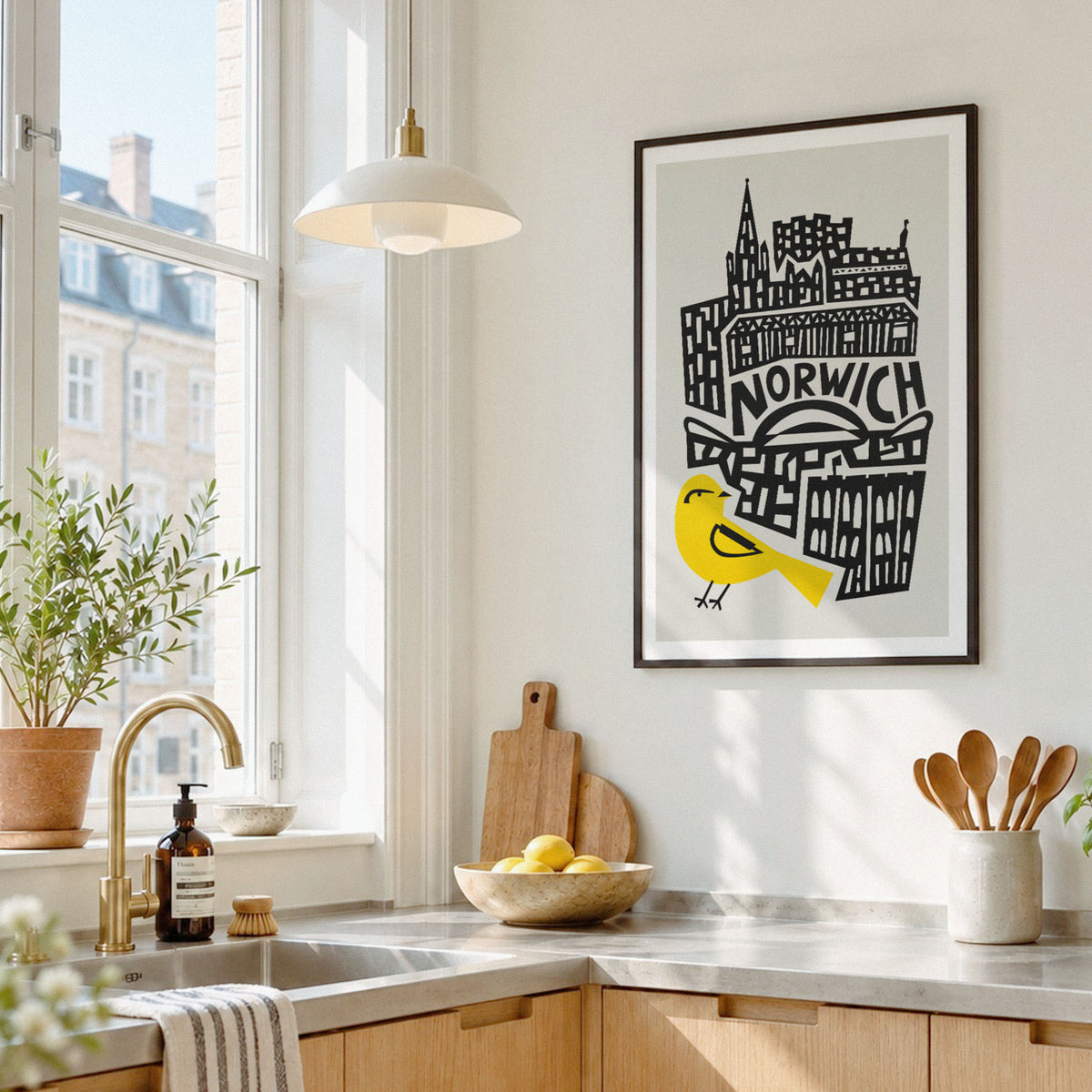 A framed art print featuring a black and white illustration of Norwich cityscape with a bright yellow bird, hanging on a light wall in a modern kitchen setting.