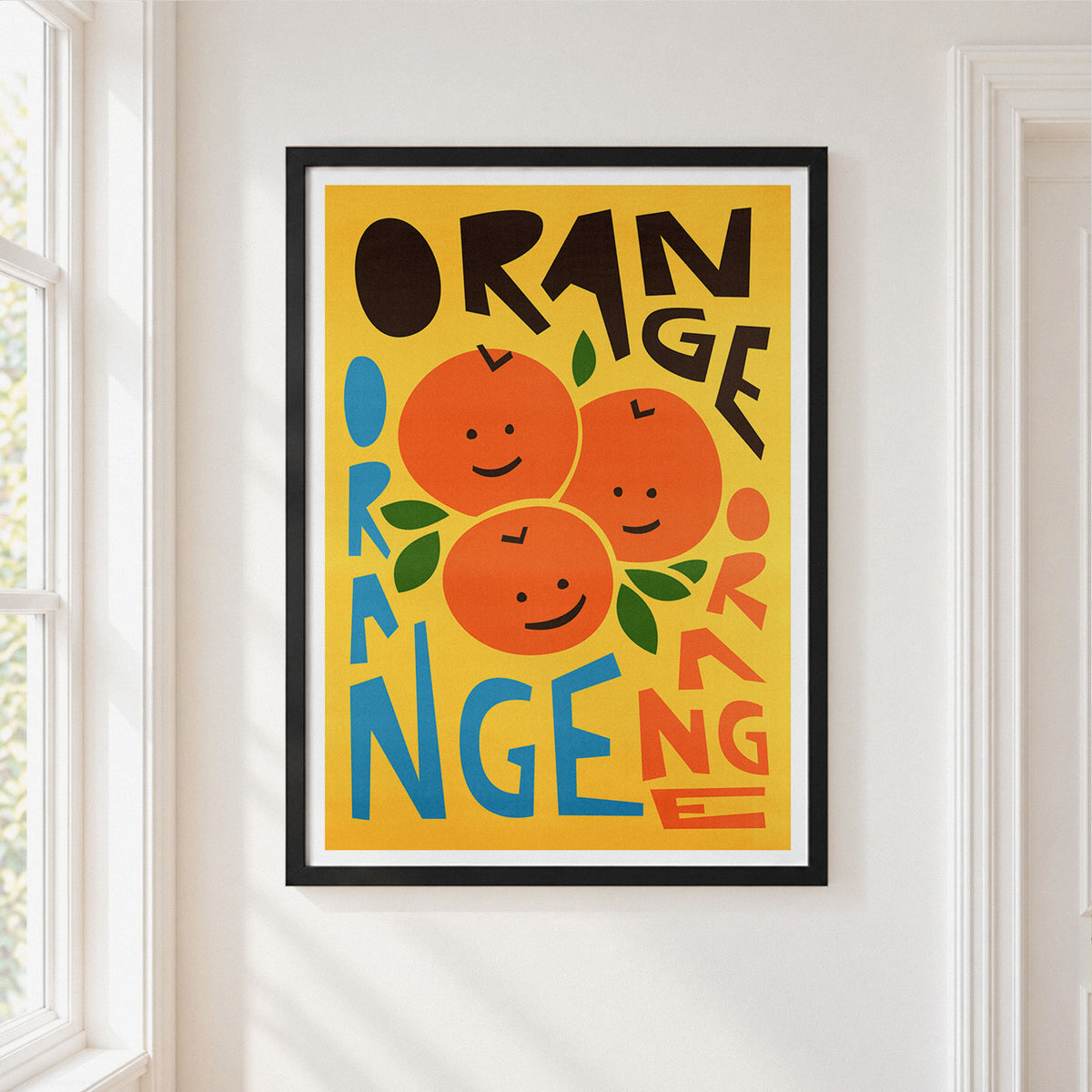 A framed art print featuring a playful orange-themed design with three smiling oranges and the word 'ORANGE' repeated in bold black, blue, and orange lettering on a mustard yellow background. The print is displayed on a white wall next to a window.