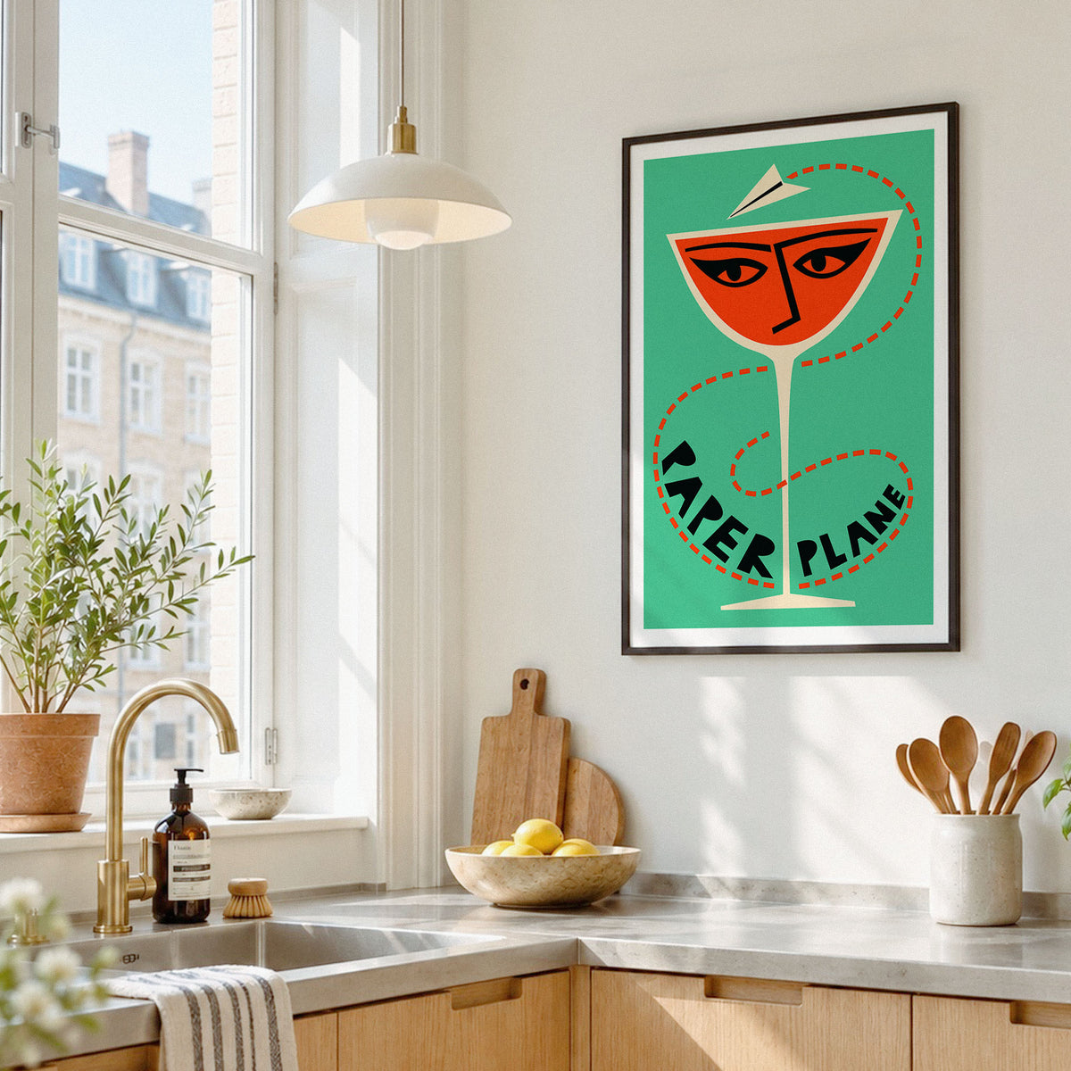 A framed pop-art style poster featuring a yellow mayonnaise can with red, black, and white typography, hanging on a white kitchen wall above a countertop with a sink, plants, and utensils.