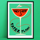 A bold paper plane cocktail print featuring a vivid orange drink in a coupe glass, illustrated with a stylised face and topped with a flying paper plane, all set against a mint green backdrop.