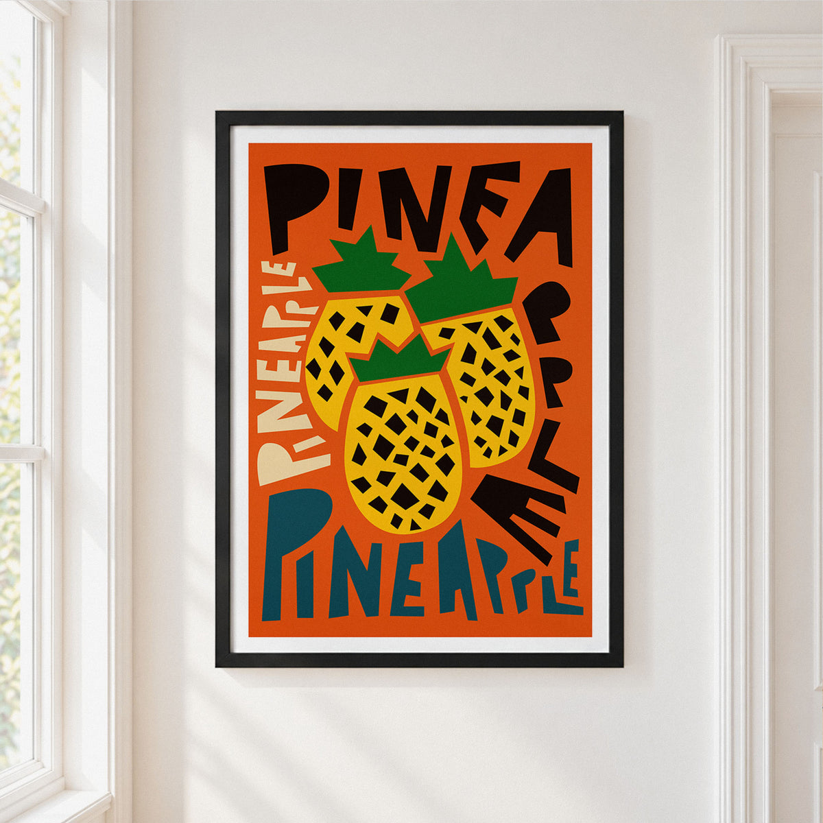 A framed art print featuring a stylized illustration of three pineapples on a bright orange background, hung on a white wall next to a window.