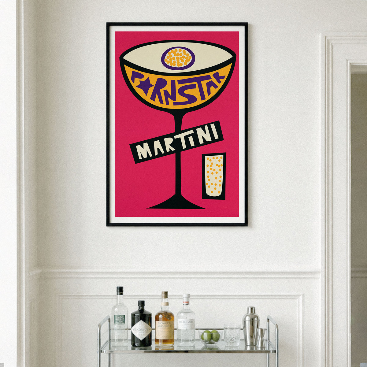 A framed pop-art style print of a martini cocktail with the word 'martini' on the glass, hanging on a white wall above a bar cart with bottles and glasses.
