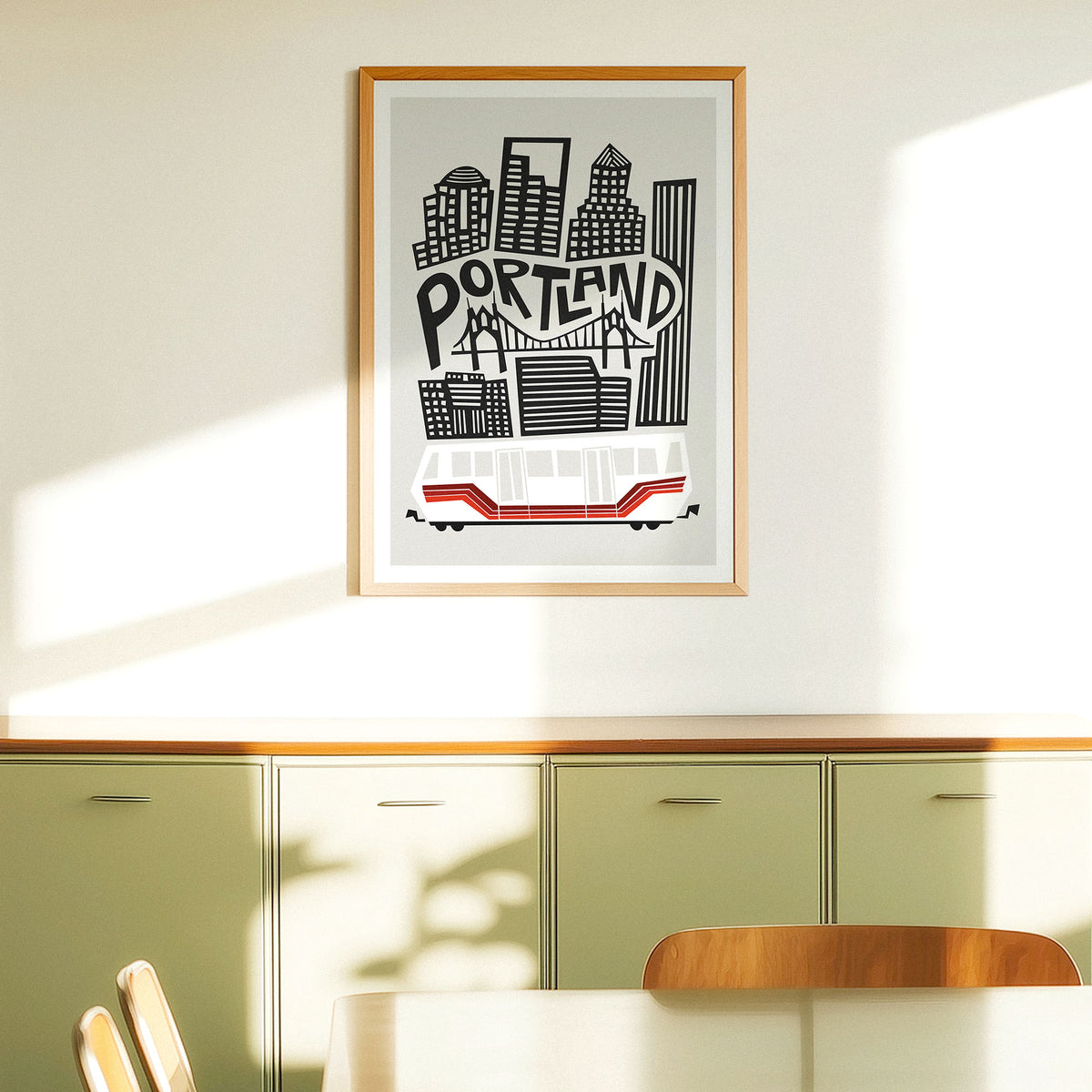 A framed art print featuring a stylized black and white Portland skyline with a red cable car, hanging on a cream wall above a green cabinet in a sunlit room.