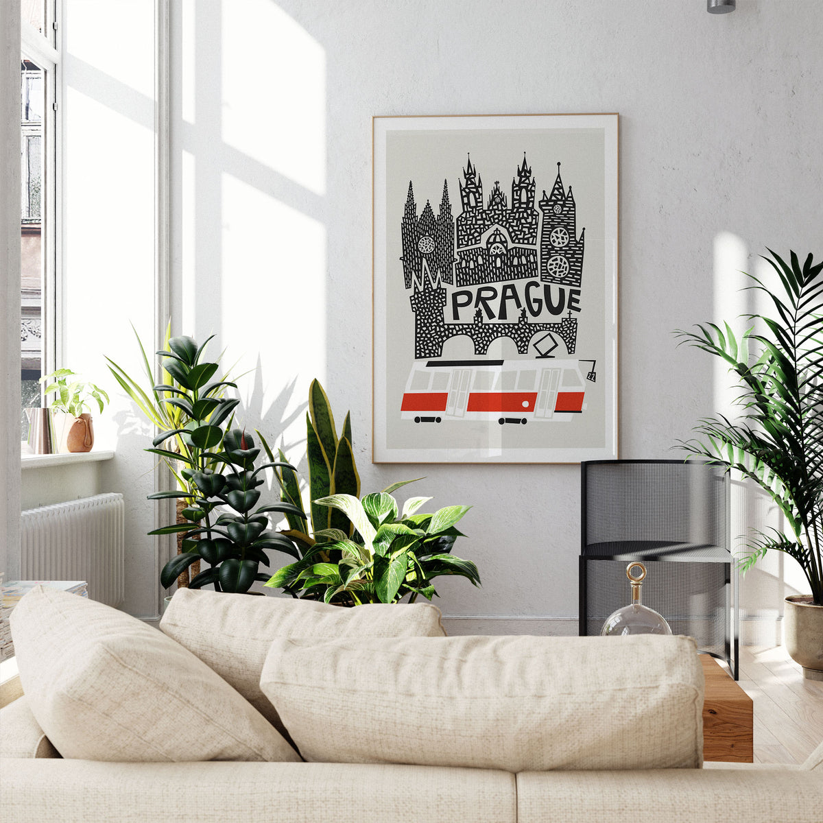 A framed art print featuring a black and white hand-drawn illustration of Prague's skyline with red accents, displayed on a light gray wall in a modern living room setting with a beige sofa and potted plants.