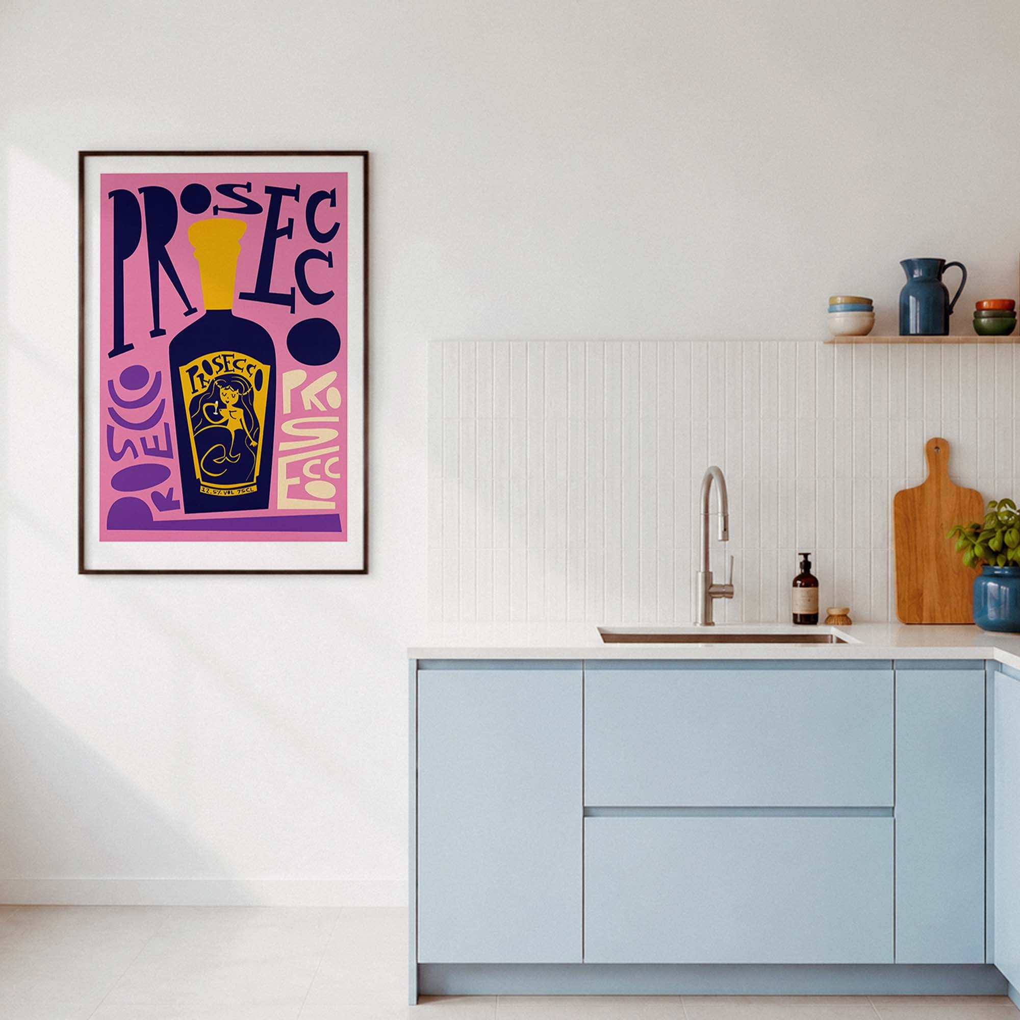 A framed pop-art poster featuring a purple and yellow bottle of prosecco illustration with stylized text, displayed on a white kitchen wall above light blue cabinets.