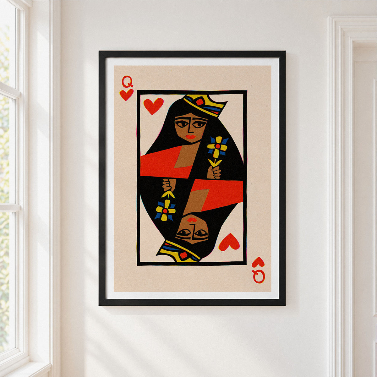 A framed art print of a stylized Queen of Hearts playing card, featuring a beige background, bold black and red geometric patterns, and red hearts, hung on a white wall next to a window.