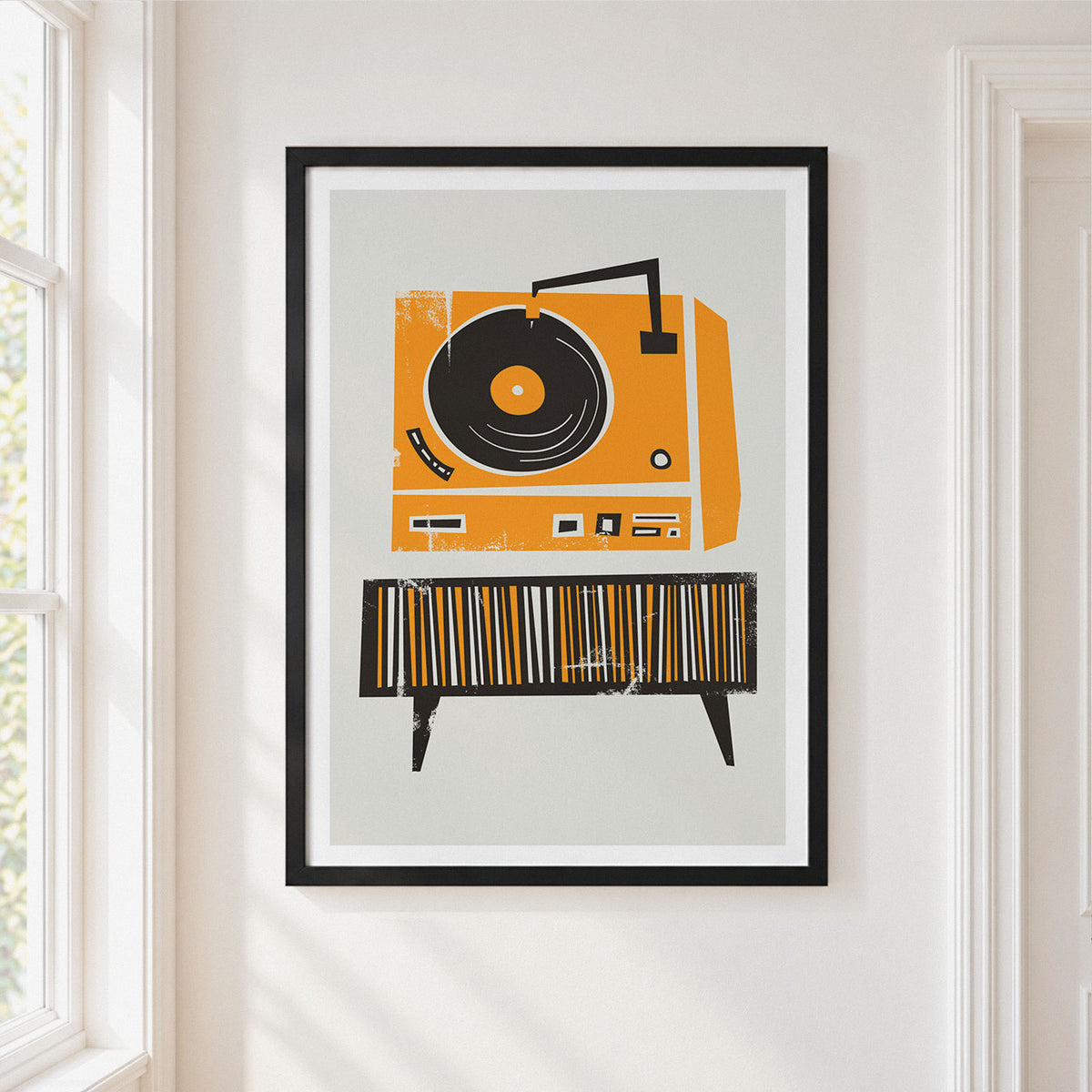 A framed art print featuring a retro orange and black vinyl record player on a striped cabinet, displayed on a white wall in a bright room next to a window.