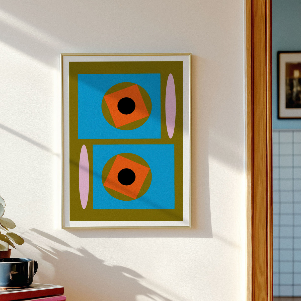 A framed abstract geometric art print hangs on a white wall. The print features two stacked blue rectangles with orange squares, black circles, olive green shapes, and pink vertical ovals.