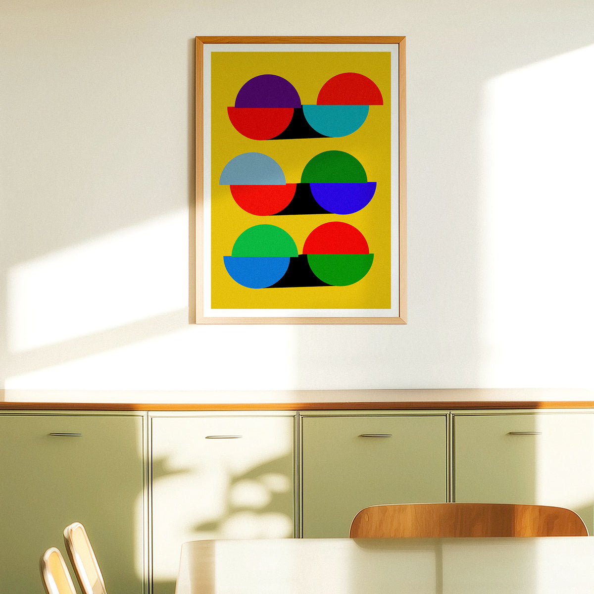 A framed abstract art print featuring colorful geometric circles on a yellow background, hanging on a white wall above a green cabinet in a modern room.