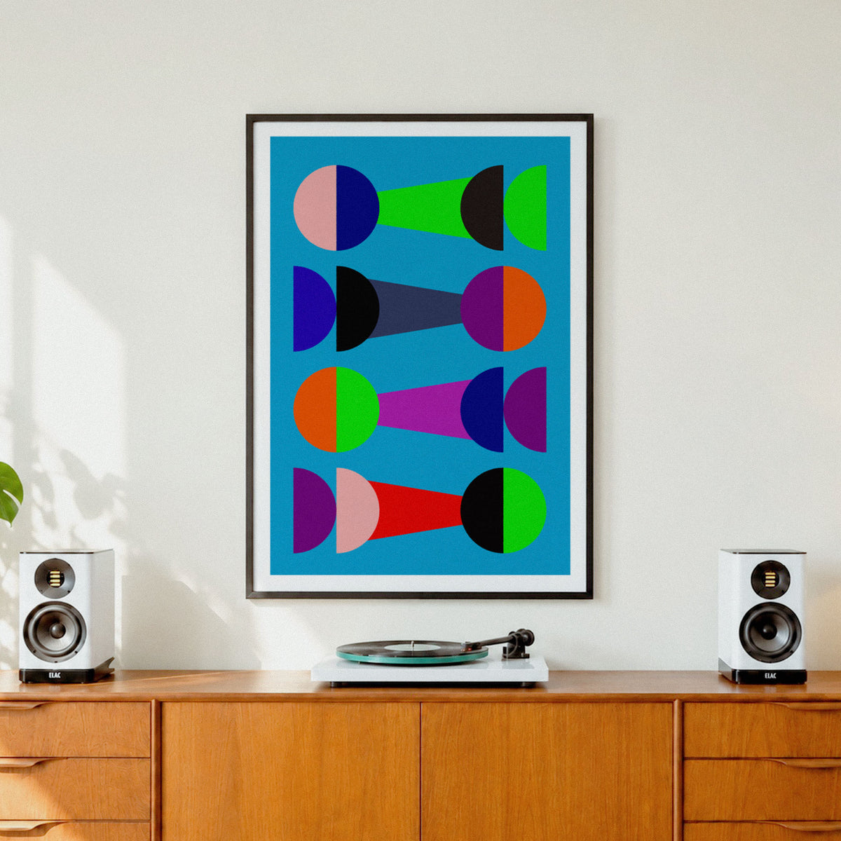 A framed abstract geometric art print with colorful semicircles and shapes on a bright blue background, hanging on a white wall above a wooden credenza with speakers and a turntable.
