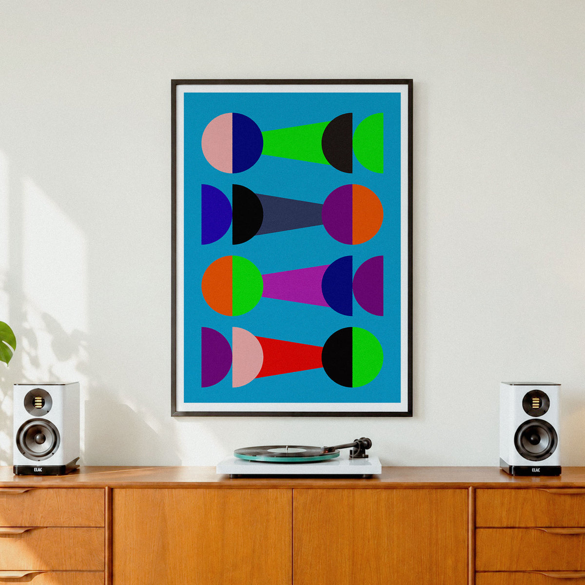 A framed abstract geometric art print with colorful semicircles and shapes on a bright blue background, hanging on a white wall above a wooden credenza with speakers and a turntable.