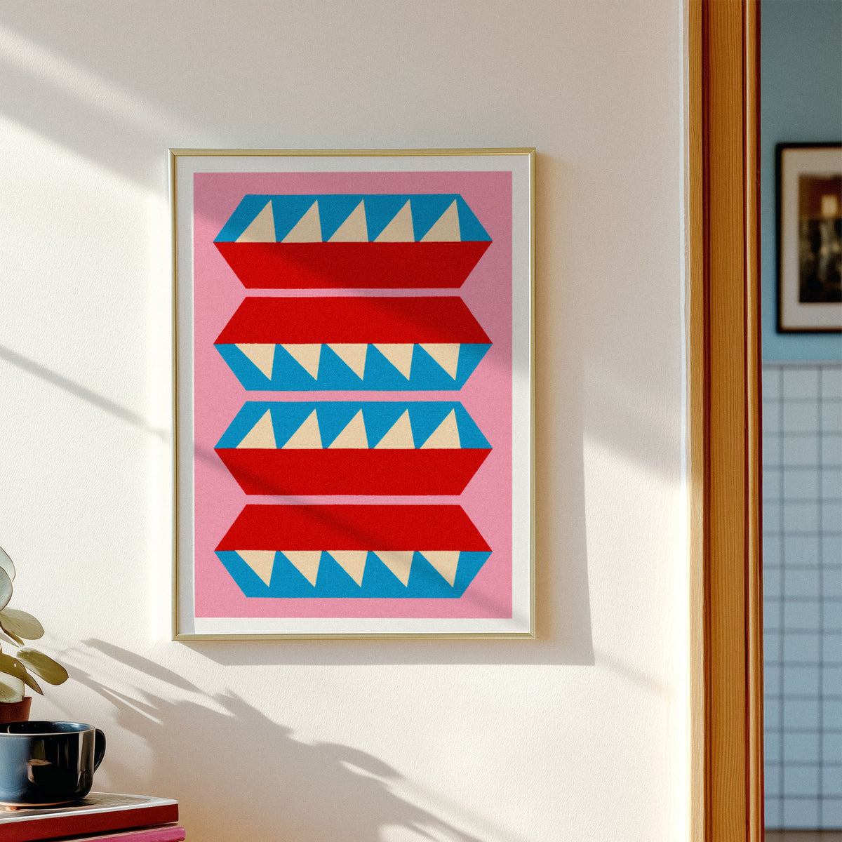 A framed art print featuring a mid-century modern geometric pattern of red and blue stylized shapes with cream triangles on a pink background, hung on a white wall next to a window.
