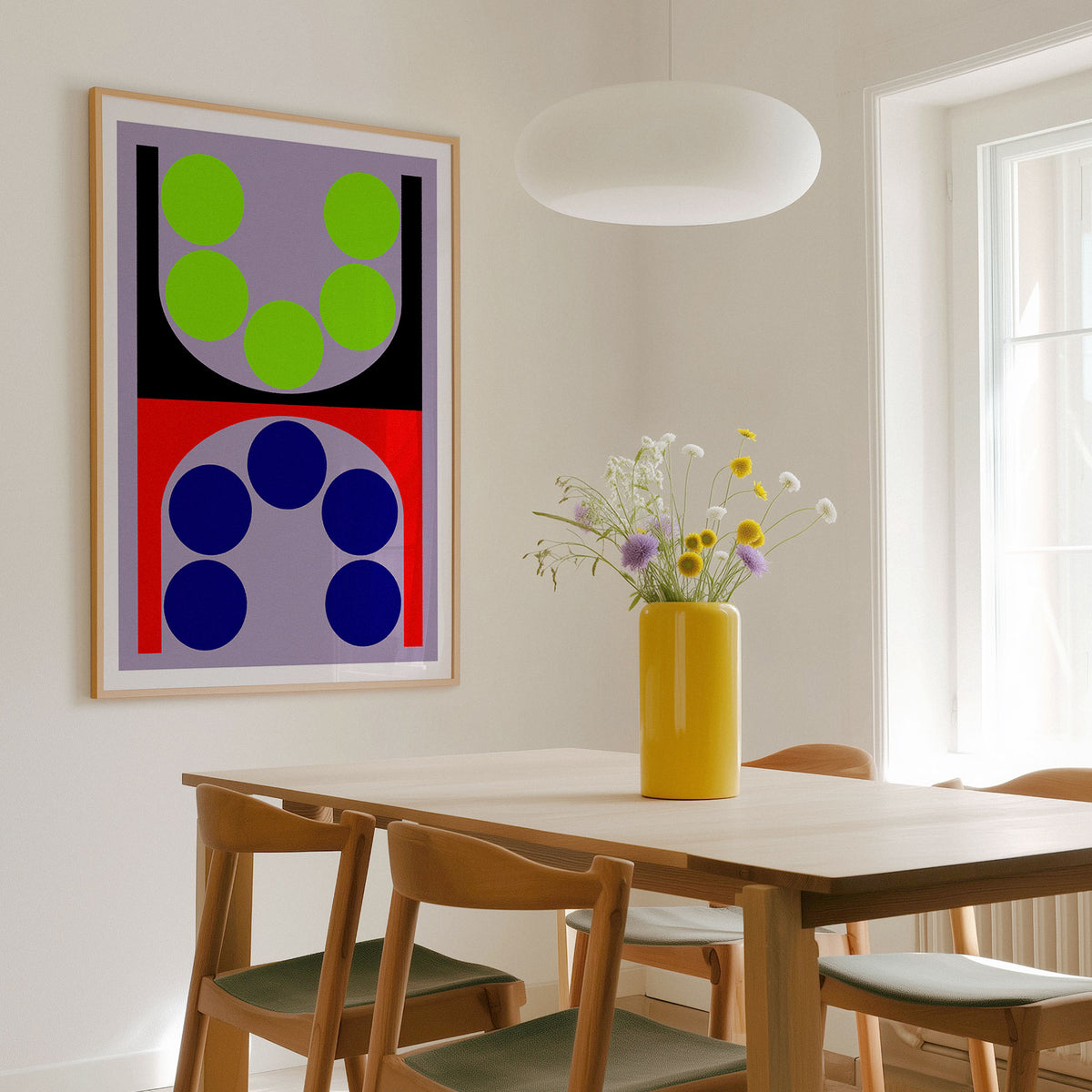 A framed abstract geometric wall art print hangs on a white dining room wall above a wooden table with chairs and a yellow vase of flowers. The artwork features bold blocks of red, purple, black, blue, and green.