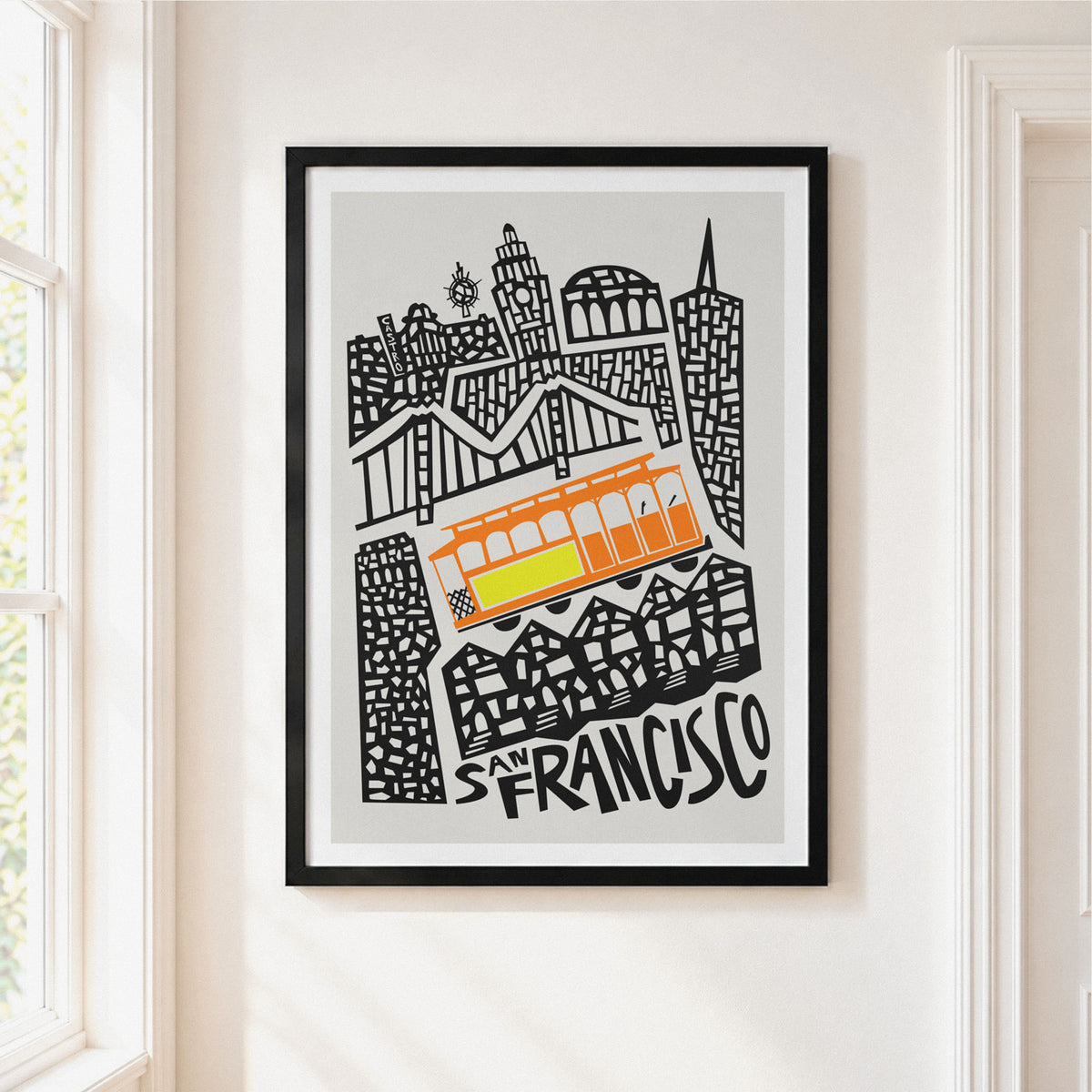 Framed San Francisco cityscape art print hanging on a white wall, featuring black line art of buildings and a bright orange and yellow tram on a light gray background.