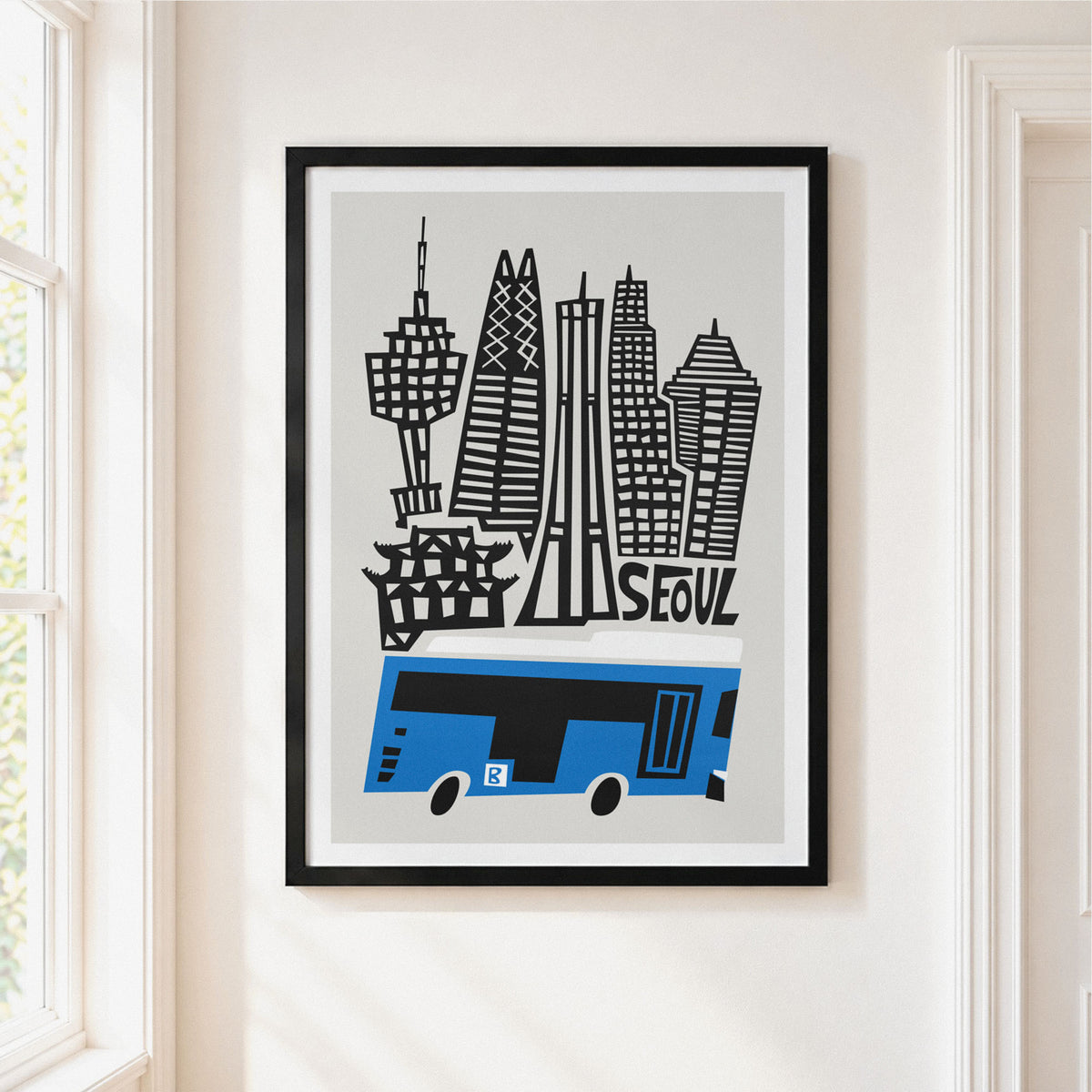 A framed art print featuring a stylized black and white Seoul skyline with a blue bus, hanging on a light beige wall in a bright room.