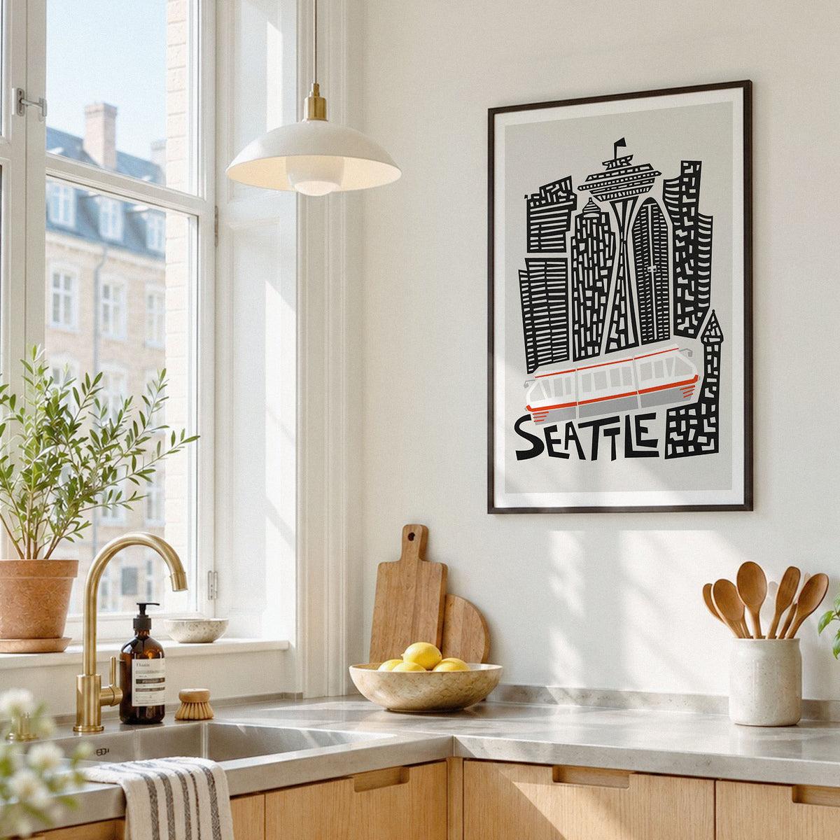 A framed Seattle cityscape art print hangs on a white kitchen wall above a countertop with a bowl of lemons, wooden cutting boards, and a hanging pendant light nearby.