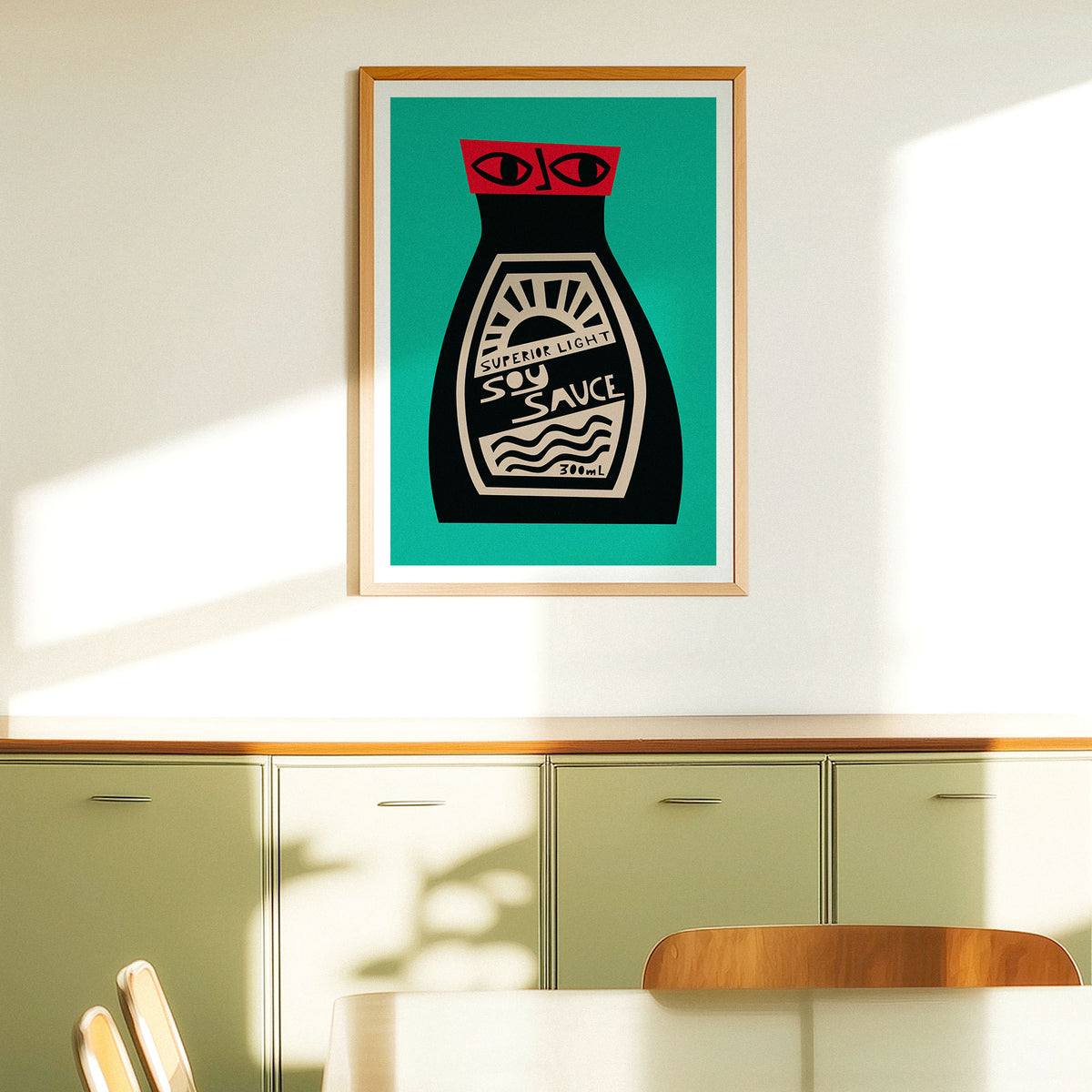 A framed art print of a retro soy sauce bottle on a teal background, hanging on a cream-colored wall above a green cabinet in a modern room.