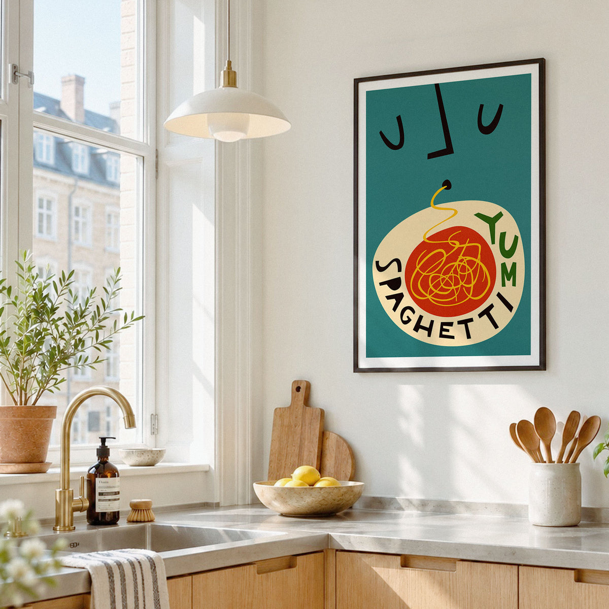 A framed art print featuring a retro-style illustration of spaghetti with the word 'SPAGHETTI' and the word 'YUM' on a teal background, hanging on a white kitchen wall above a counter with a sink, plants, and wooden utensils.