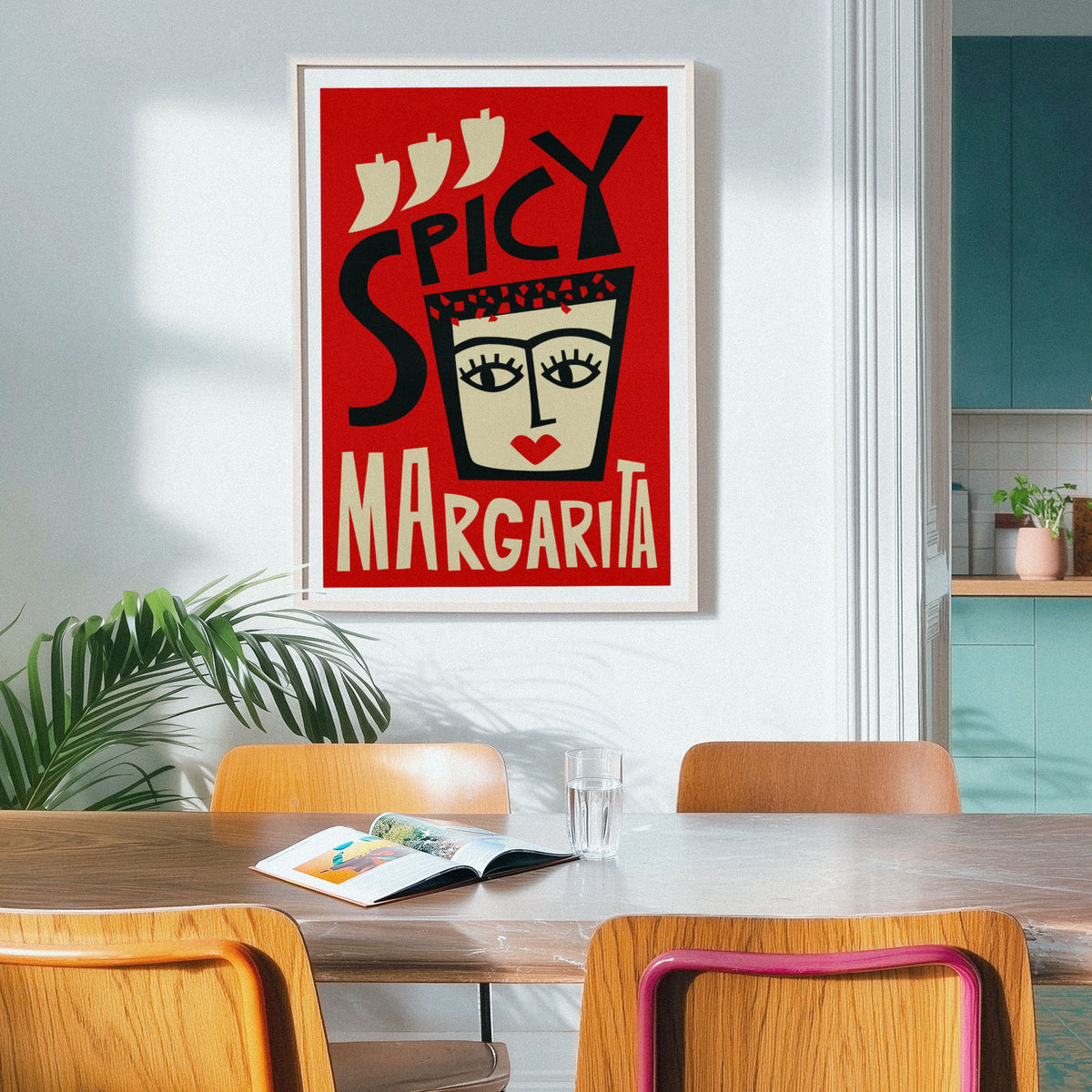 A framed art print featuring a retro-style illustration of a margarita glass with the words 'SPICY MARGARITA' on a red background, hanging on a light gray wall in a dining room setting with wooden chairs and a table.